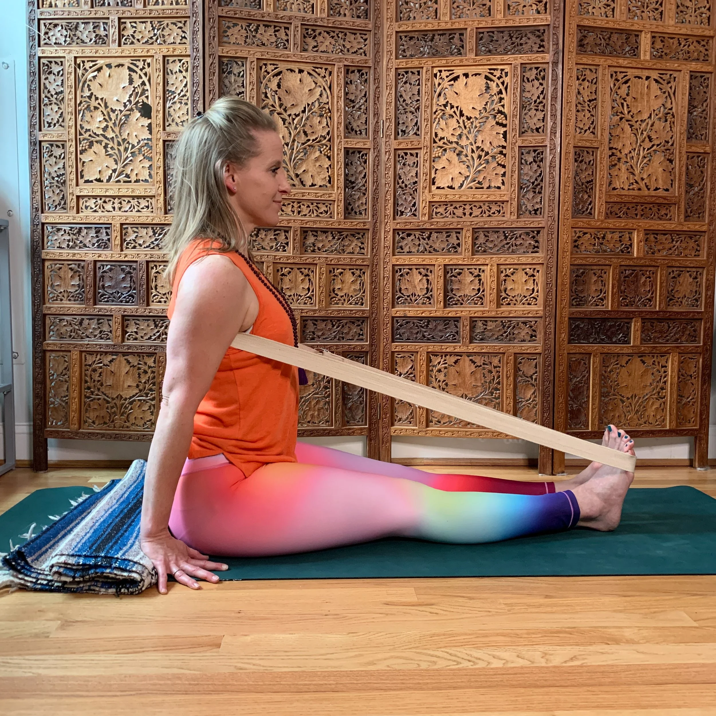 Woman sits in Staff Pose with a yoga belt around her thoracic spine and feet wearing rainbow yoga pants