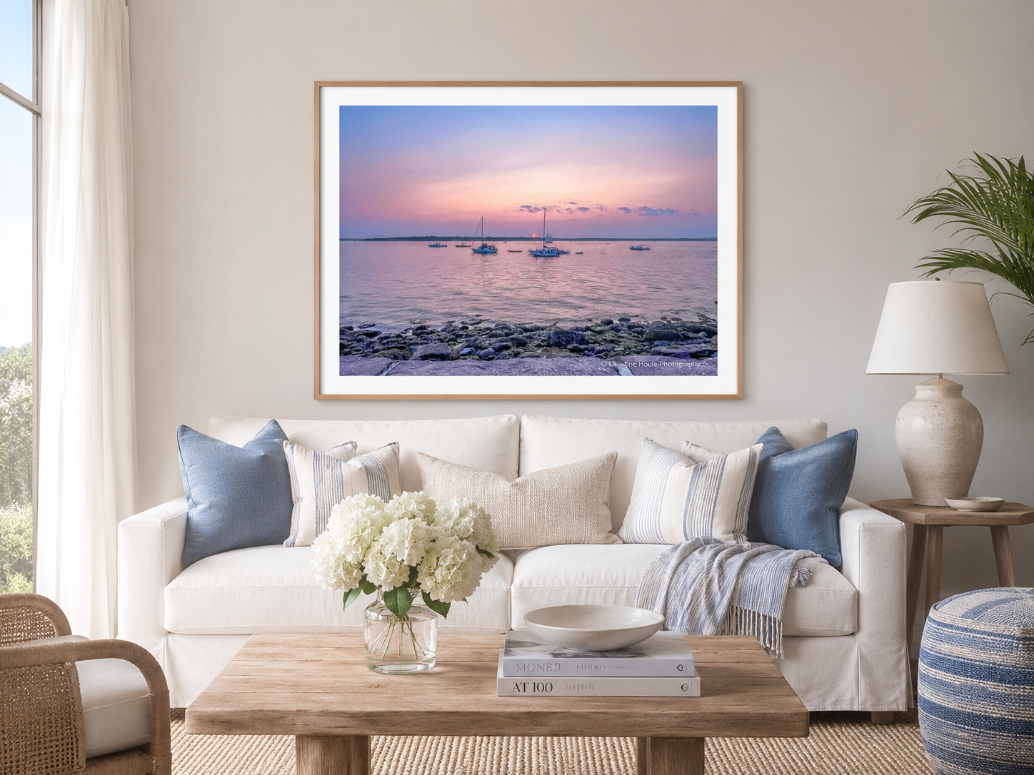 Small boats resting in a harbor beneath a lavender and rose sunset sky on the wall above a cream couch in an airy living room.