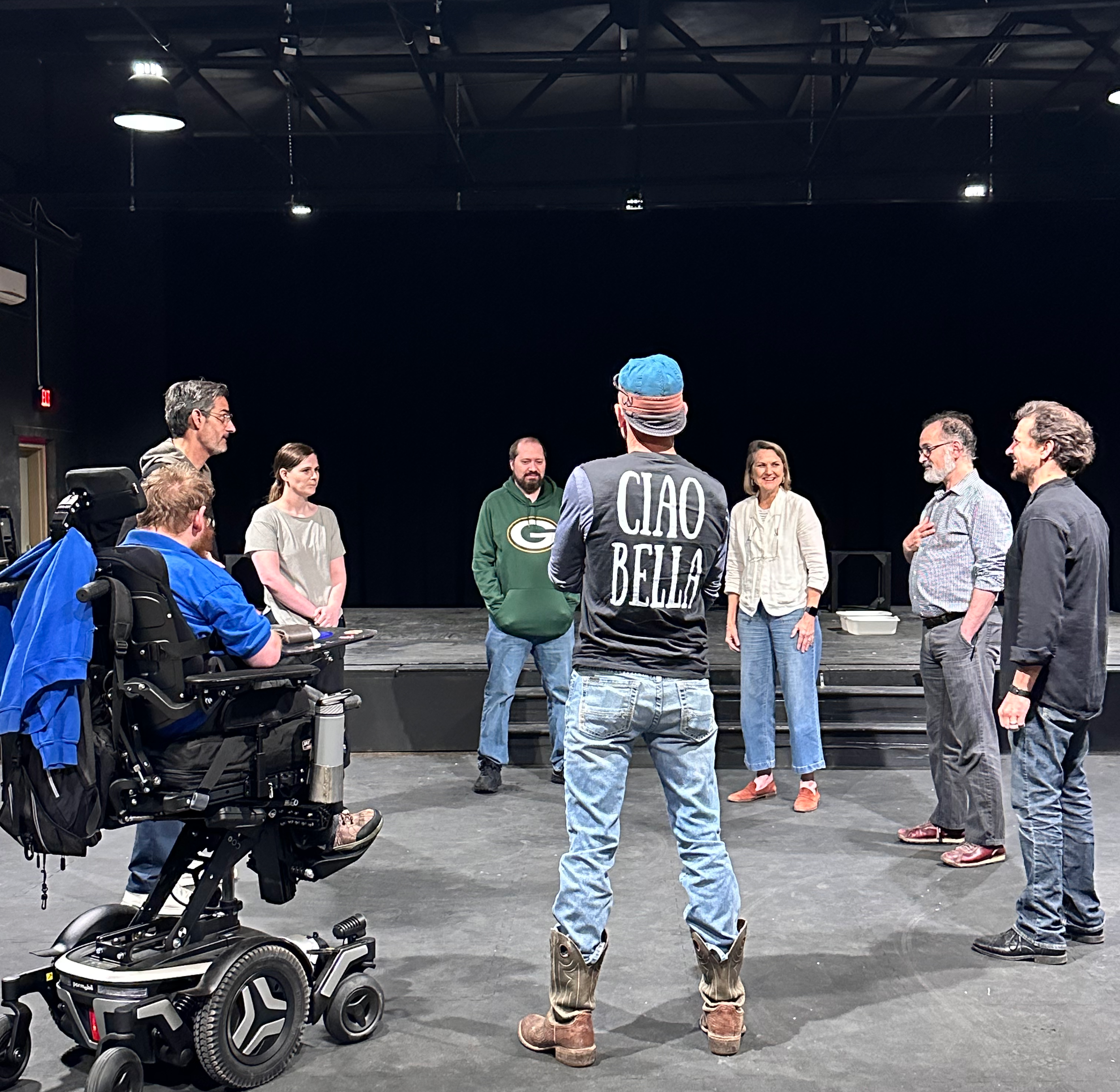 Group of people standing in a semi-circle on a stage, participating in a discussion or rehearsal, with one person in a wheelchair and a person wearing a shirt that reads 'CIAO BELLA' facing the group.