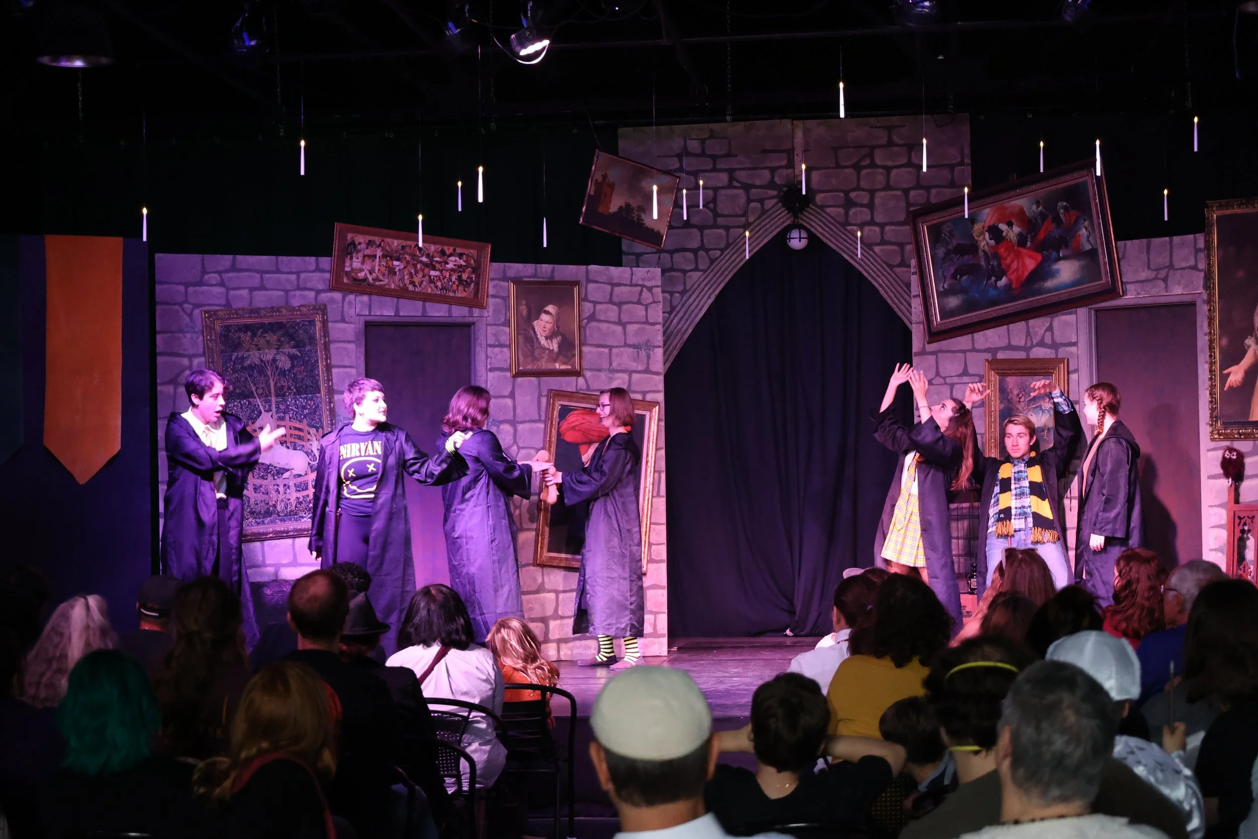 Students performing on stage in a school play, dressed in robes, with a backdrop of paintings and a castle wall, an audience seated in front watching.