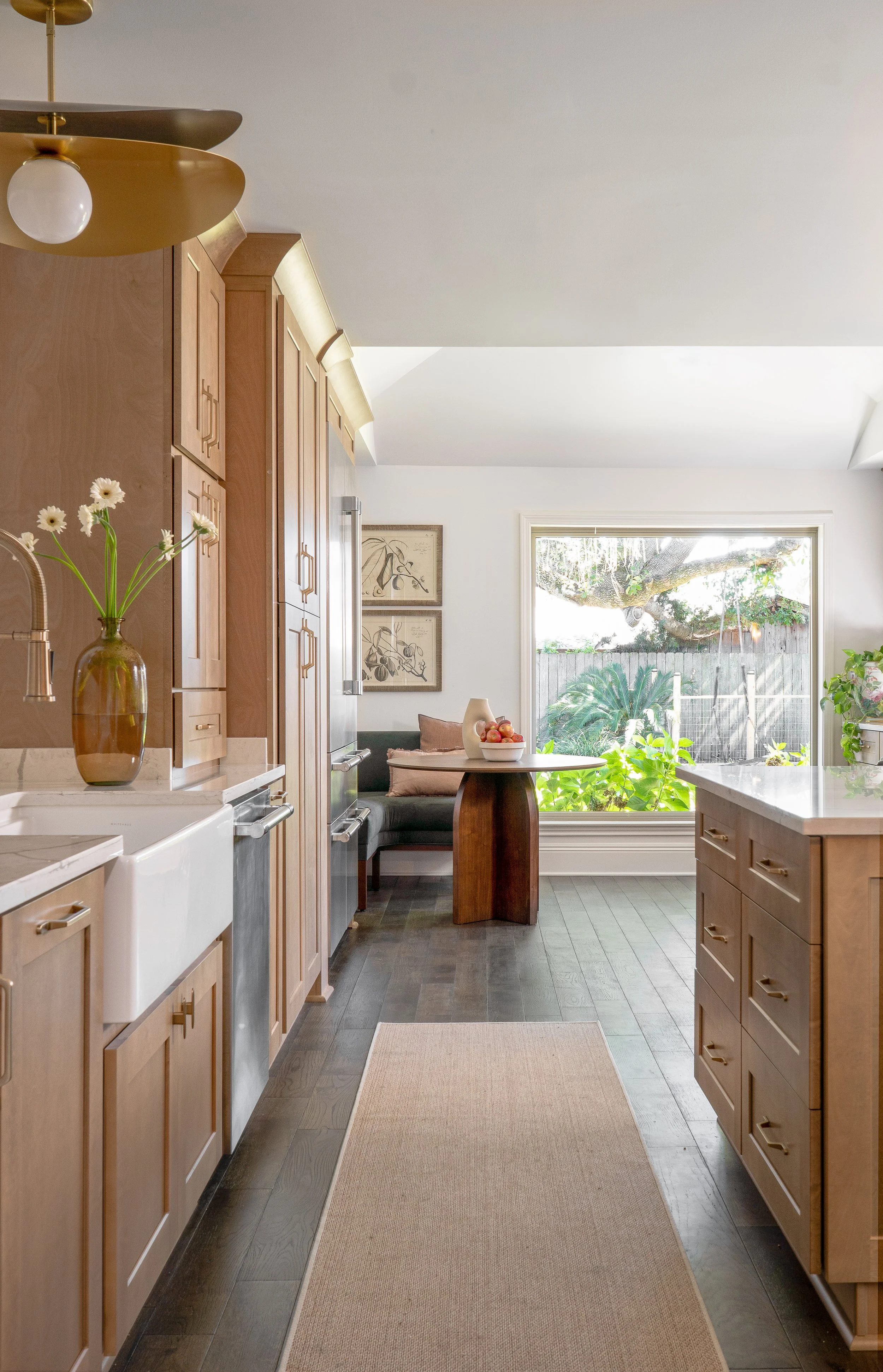 A kitchen with wooden cabinets, a white farmhouse sink, and a beige rug on dark hardwood floors. A large window shows greenery outside, and a table with a bowl of apples and decorative items is near the window.