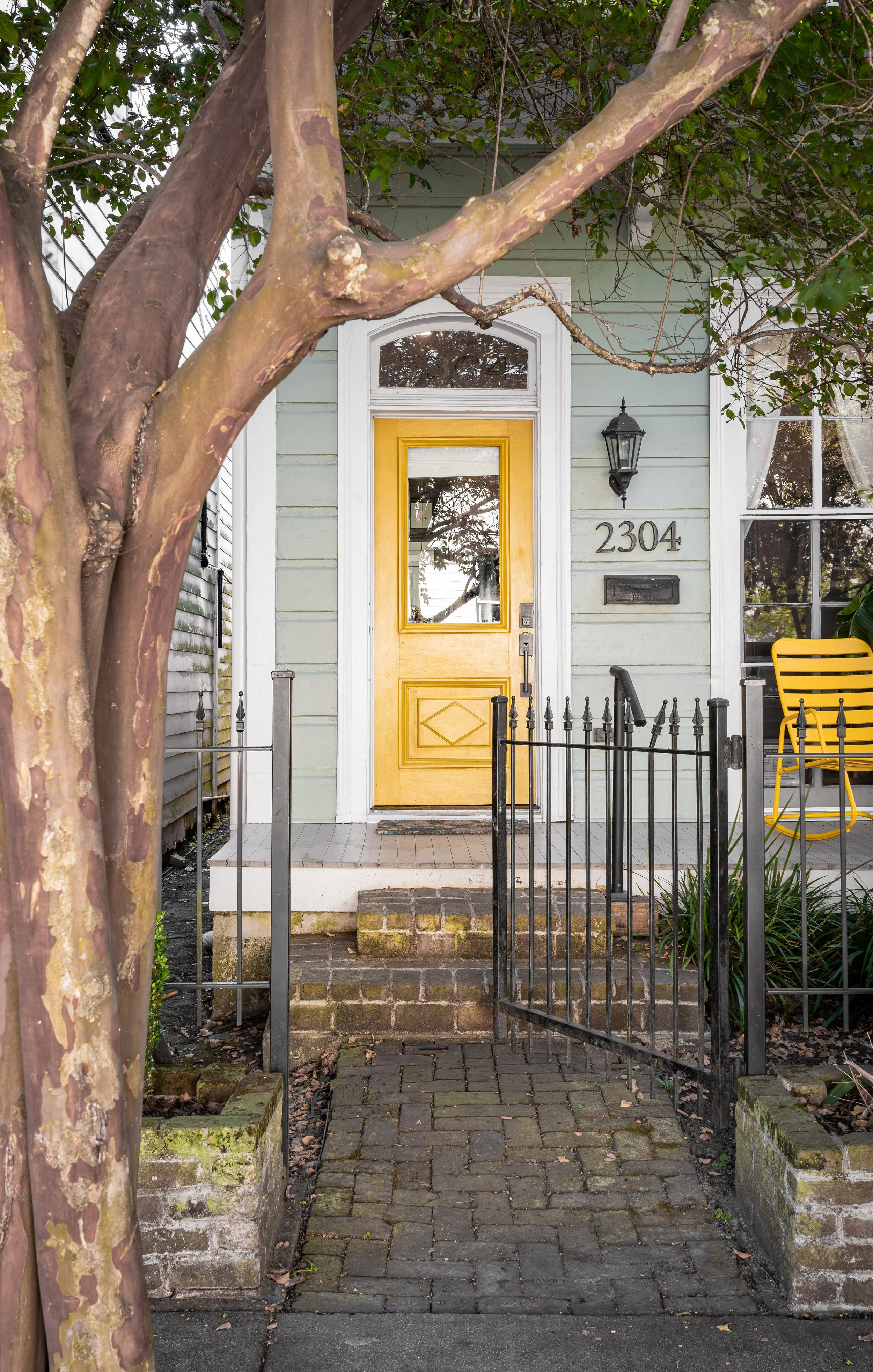 Front door of a house with a yellow door, house number 2304, two steps leading up to a small porch, a large tree with a wide trunk in front, and a black metal gate.