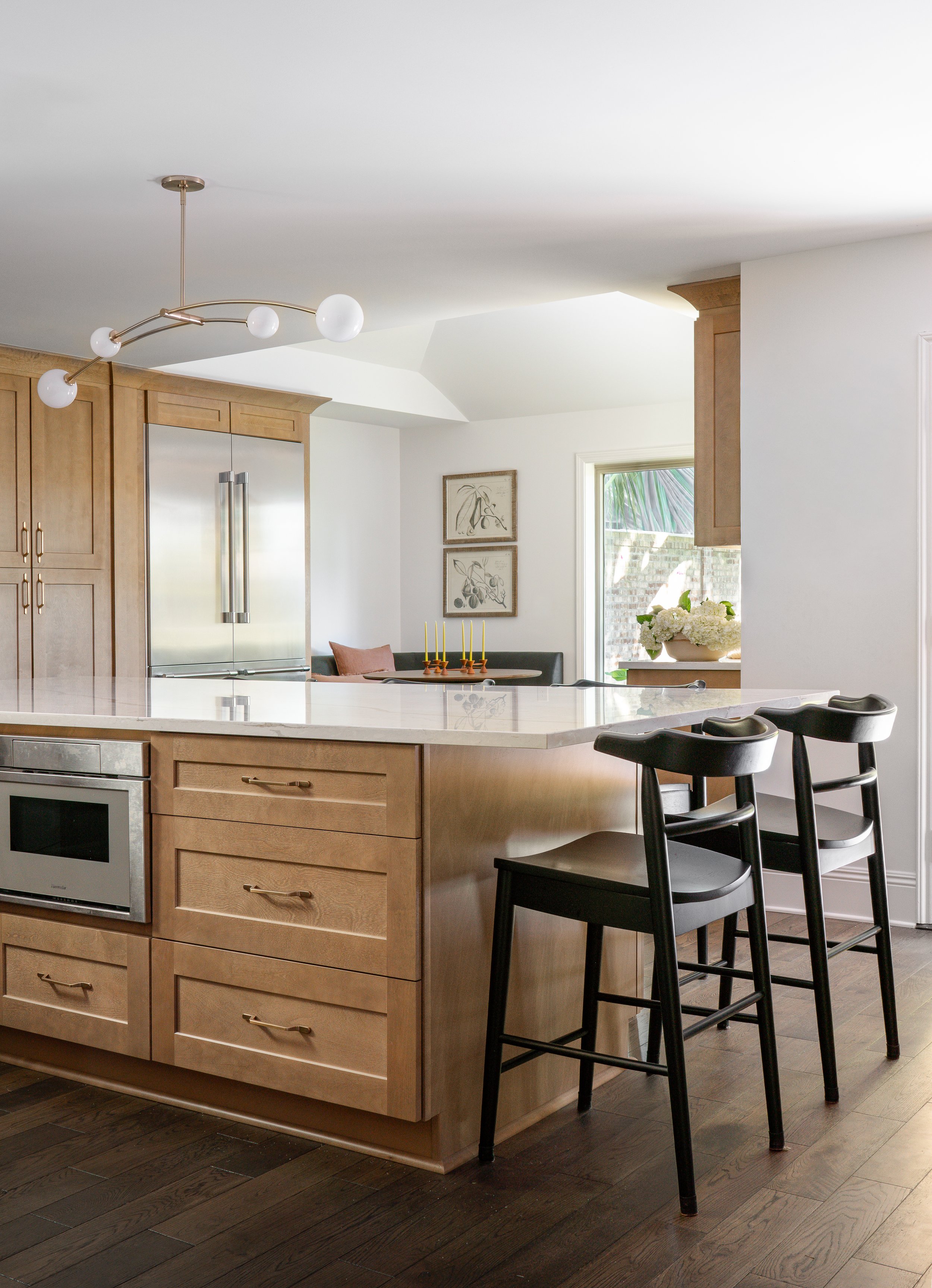 Modern kitchen with light wood cabinets, a white marble island, and black barstools. There is a stainless steel refrigerator and artwork on the wall. Natural light from a window illuminates the space.