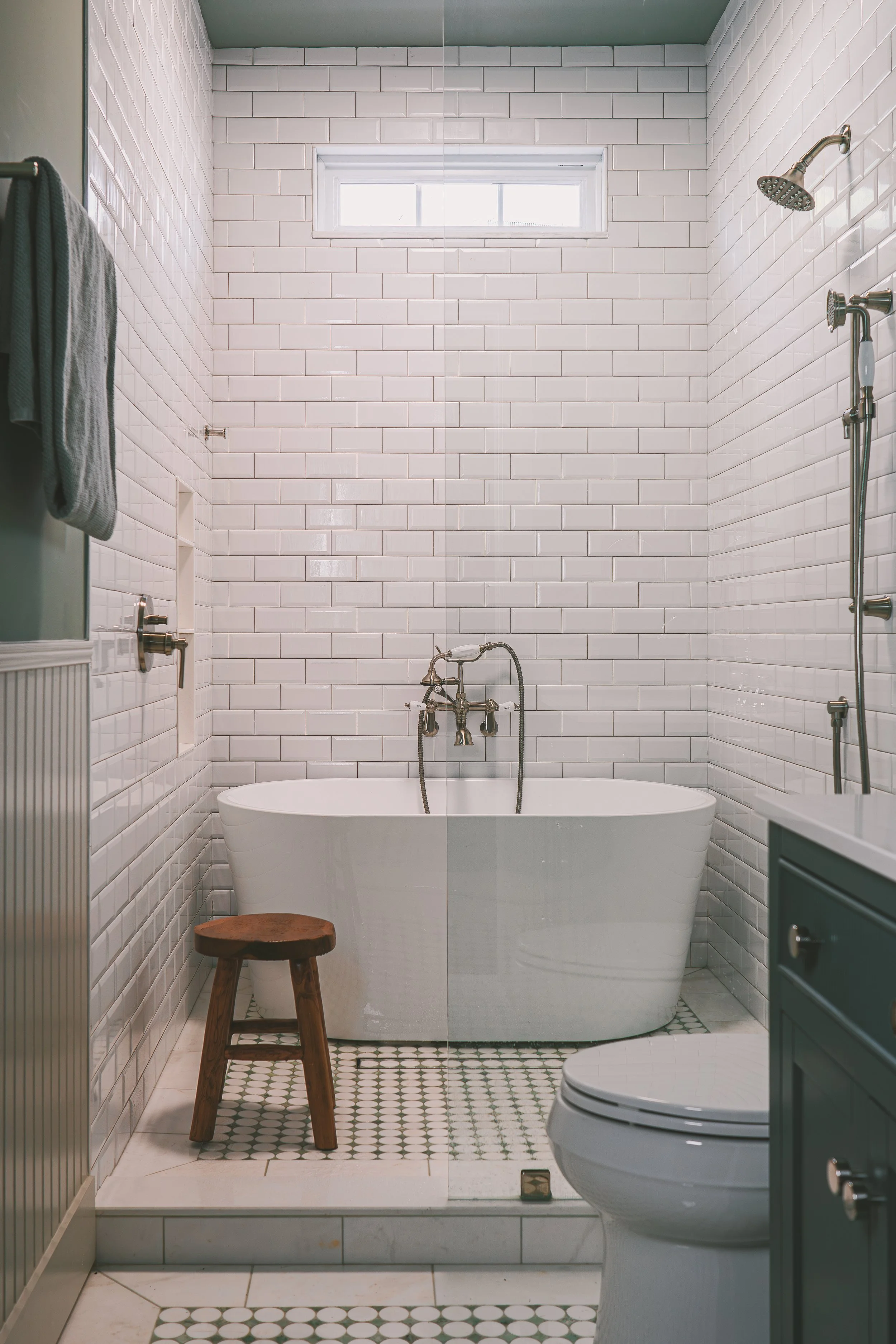 Modern bathroom with white subway tile walls, a white bathtub, a small wooden stool, a toilet, a green vanity, and a window near the ceiling.