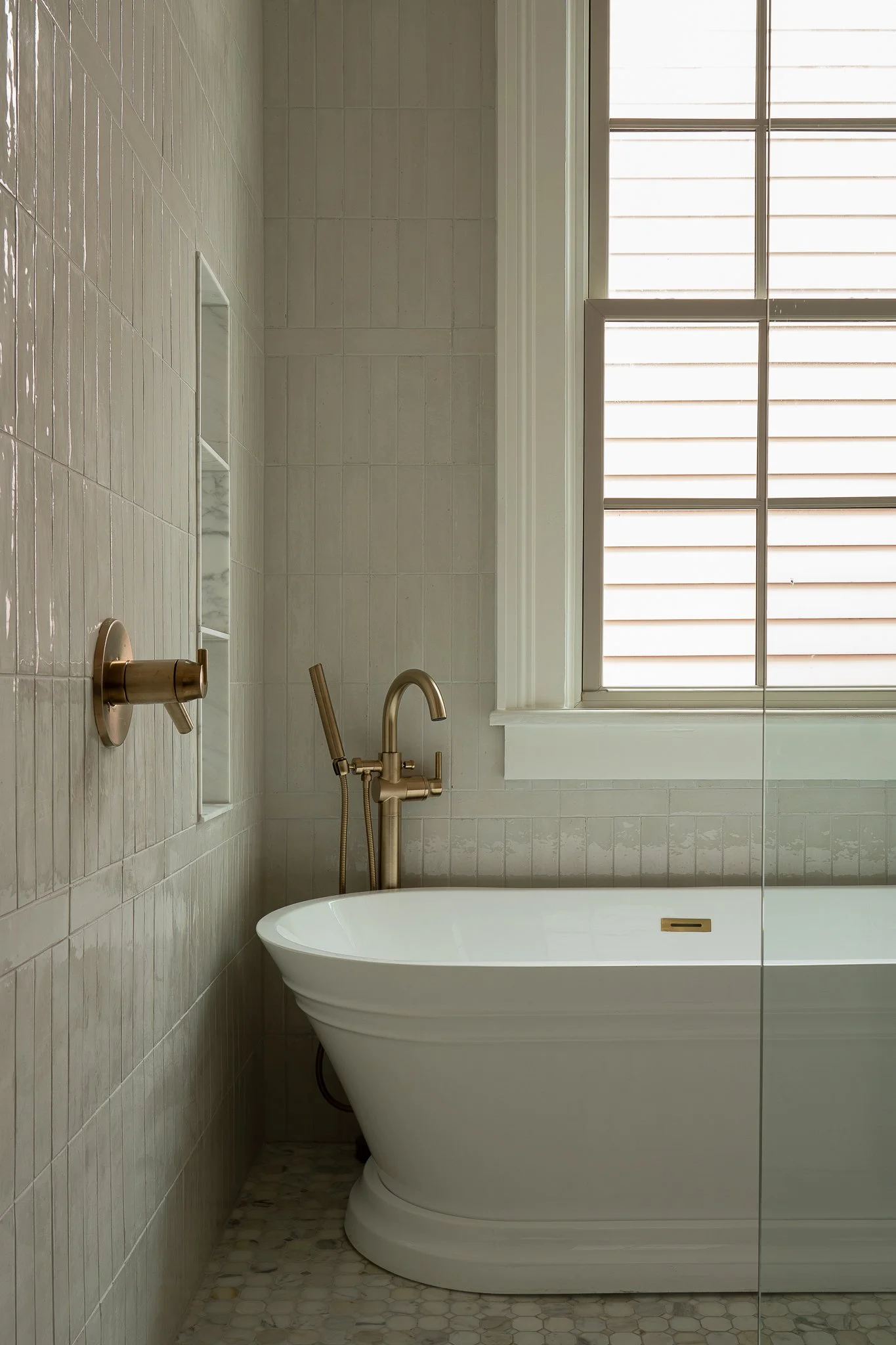 Bathroom with a white bathtub, a large window with blinds, and beige tiled walls.