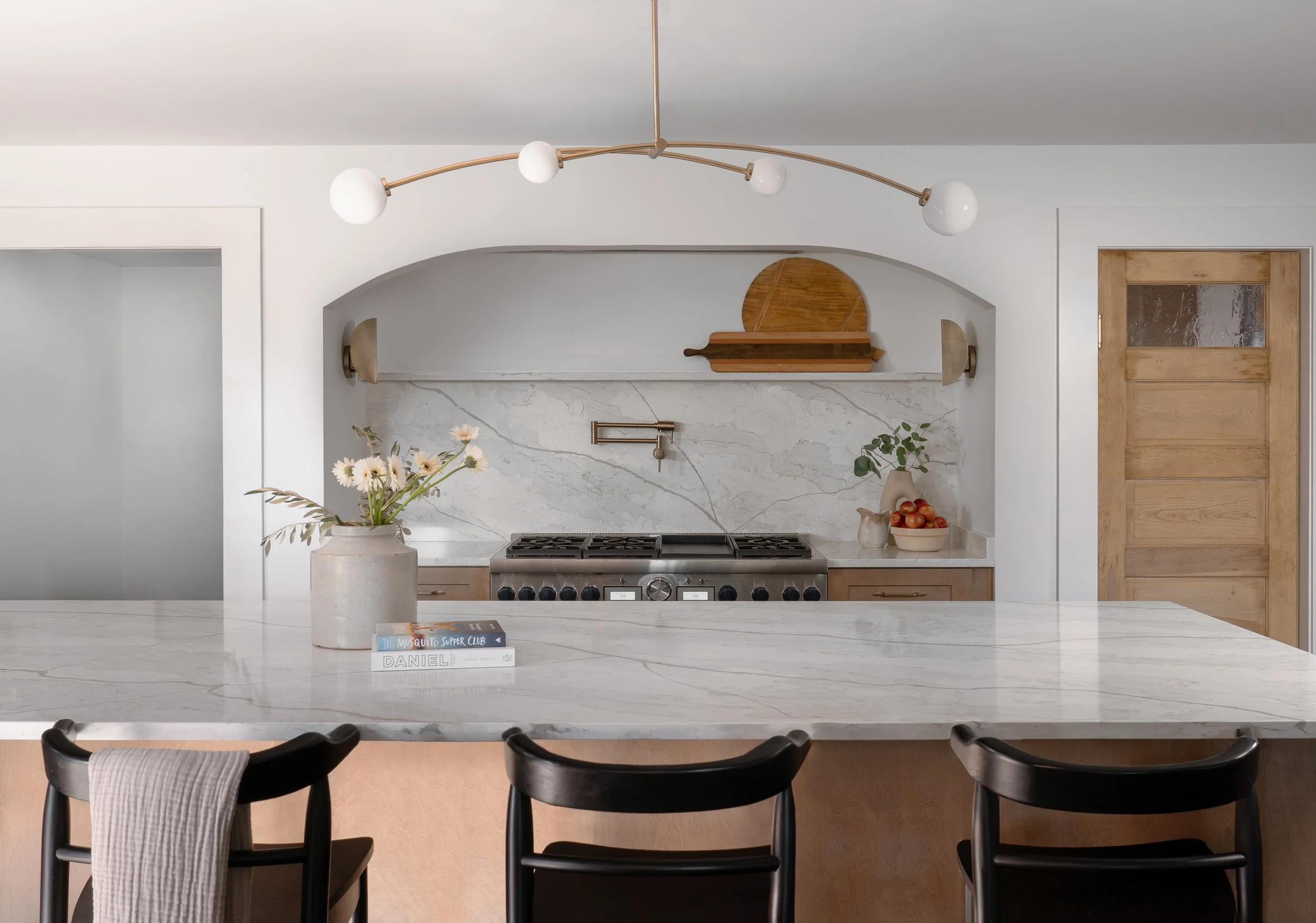 Modern kitchen with a marble island, black chairs, a vase with flowers, and a stove with a marble backsplash. There are wooden doors and shelves, with a stylish overhead light fixture.