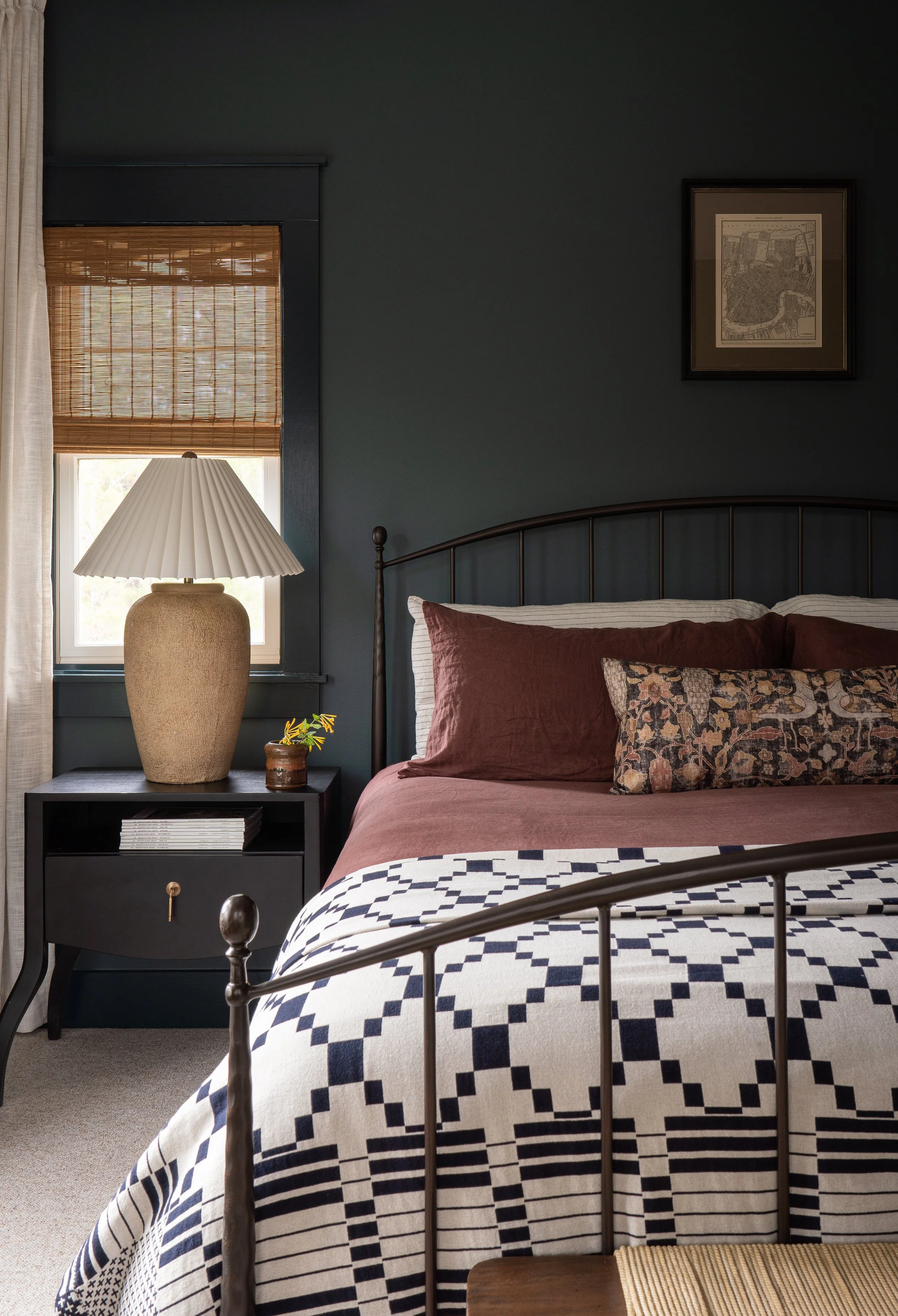 A cozy bedroom with dark teal walls, a metal bed frame, and patterned bedding. Beside the bed is a black nightstand with a large beige table lamp, a small plant, and some magazines. A window with a bamboo shade filters natural light into the room, an