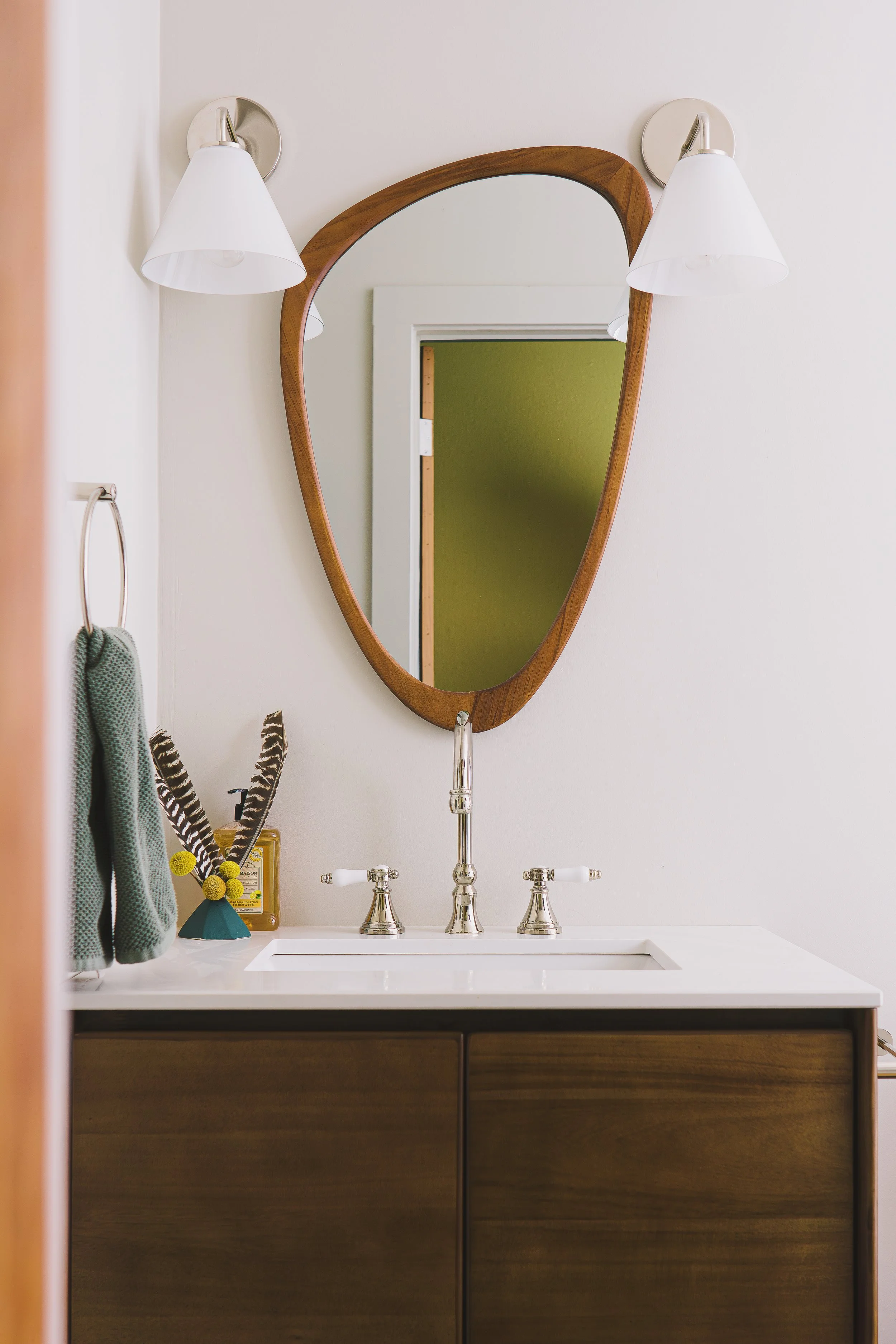 Bathroom vanity with wooden cabinet, white countertop, and silver fixtures. An oval mirror with a wooden frame is mounted above the sink. Two wall-mounted white lamps and a towel ring with a green towel are visible. Small decorative items are placed 
