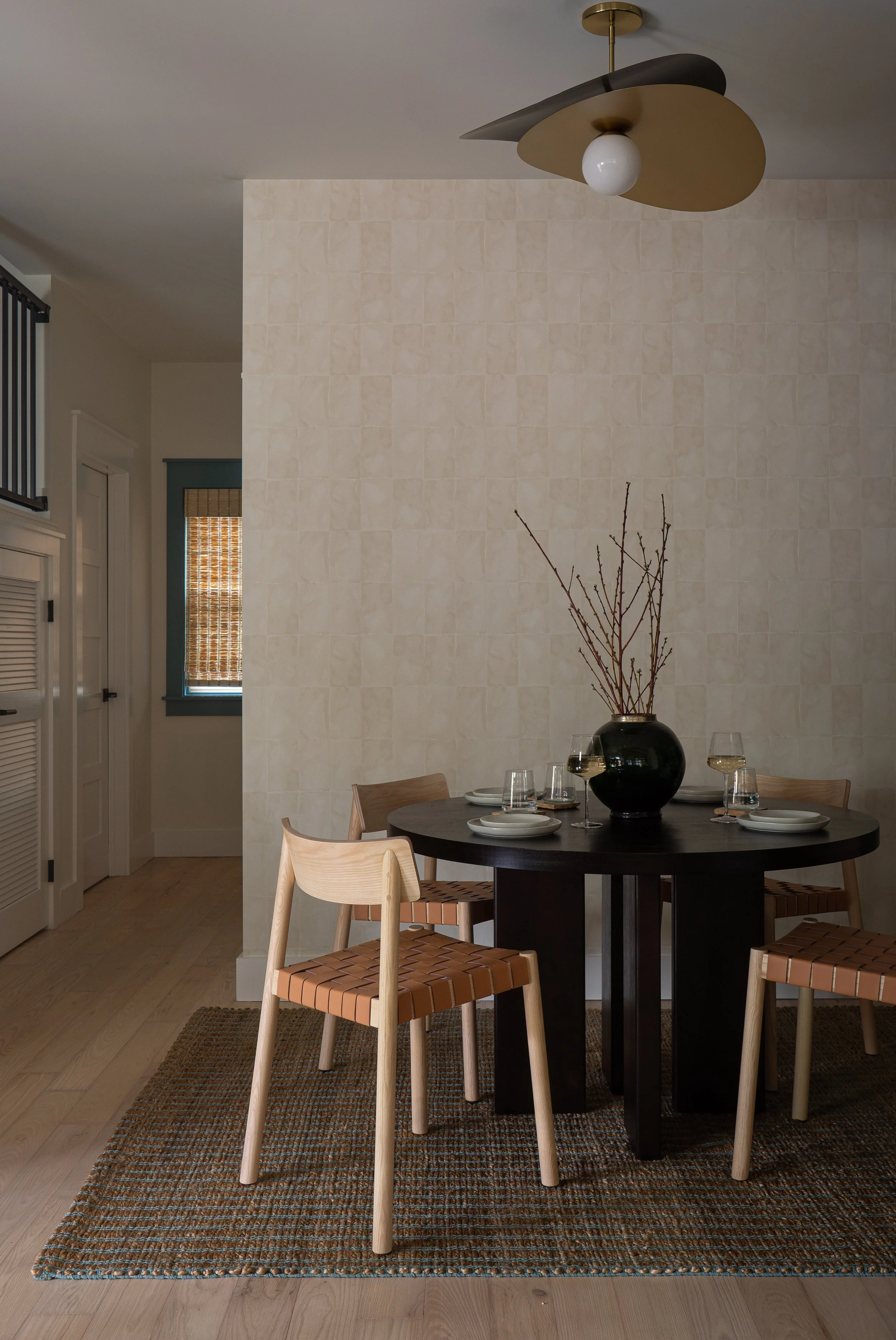 A round black dining table set with a black vase containing bare branches and four glasses of white wine, on a patterned brown rug with four wooden chairs around it, in a room with beige tiled walls and a modern ceiling light fixture.