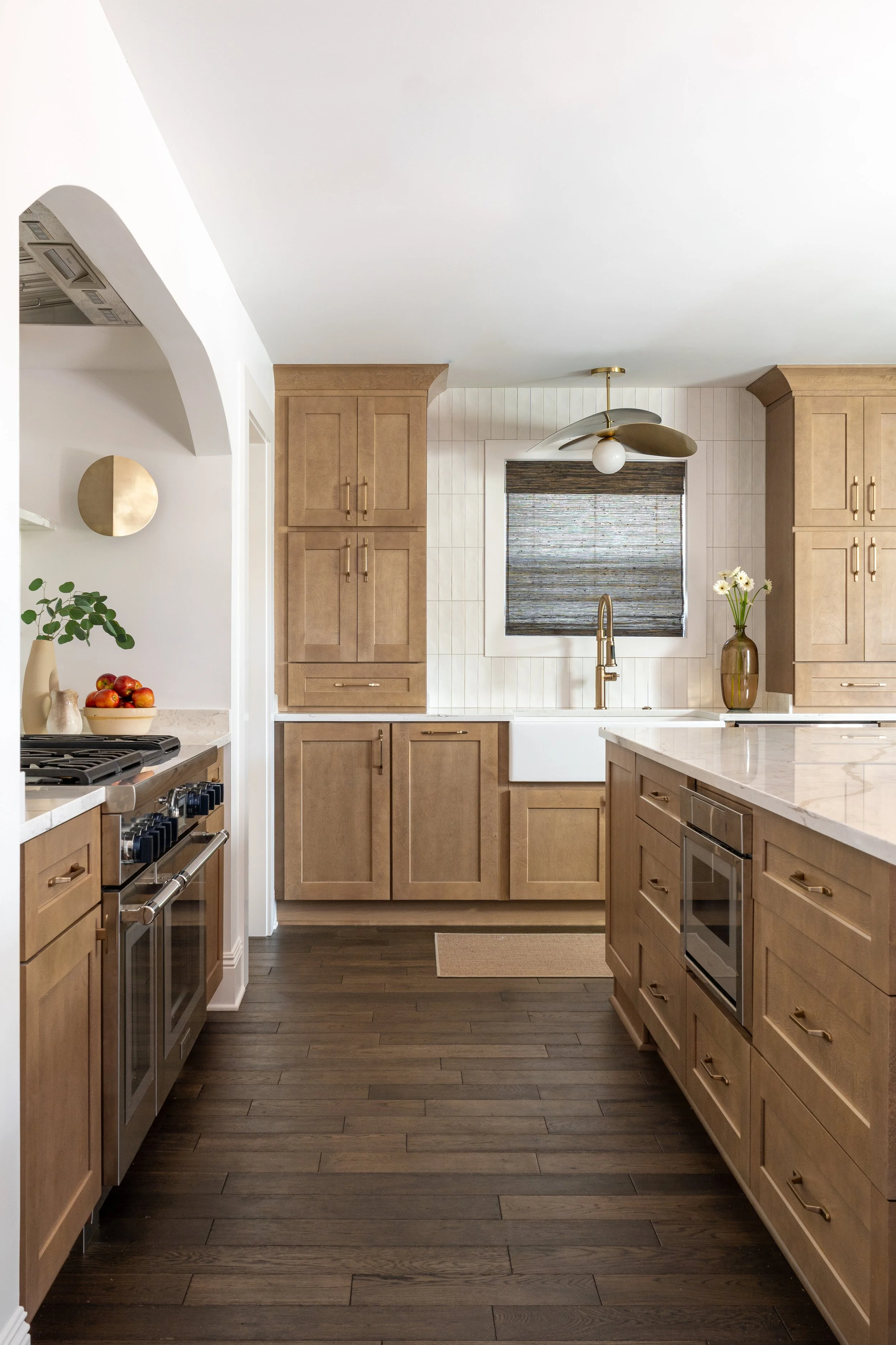 A modern kitchen with light wood cabinetry, white countertops, dark wood flooring, and a white farmhouse sink beneath a window with a dark window shade. Decor includes a potted plant, a bowl of apples, and a vase with flowers.