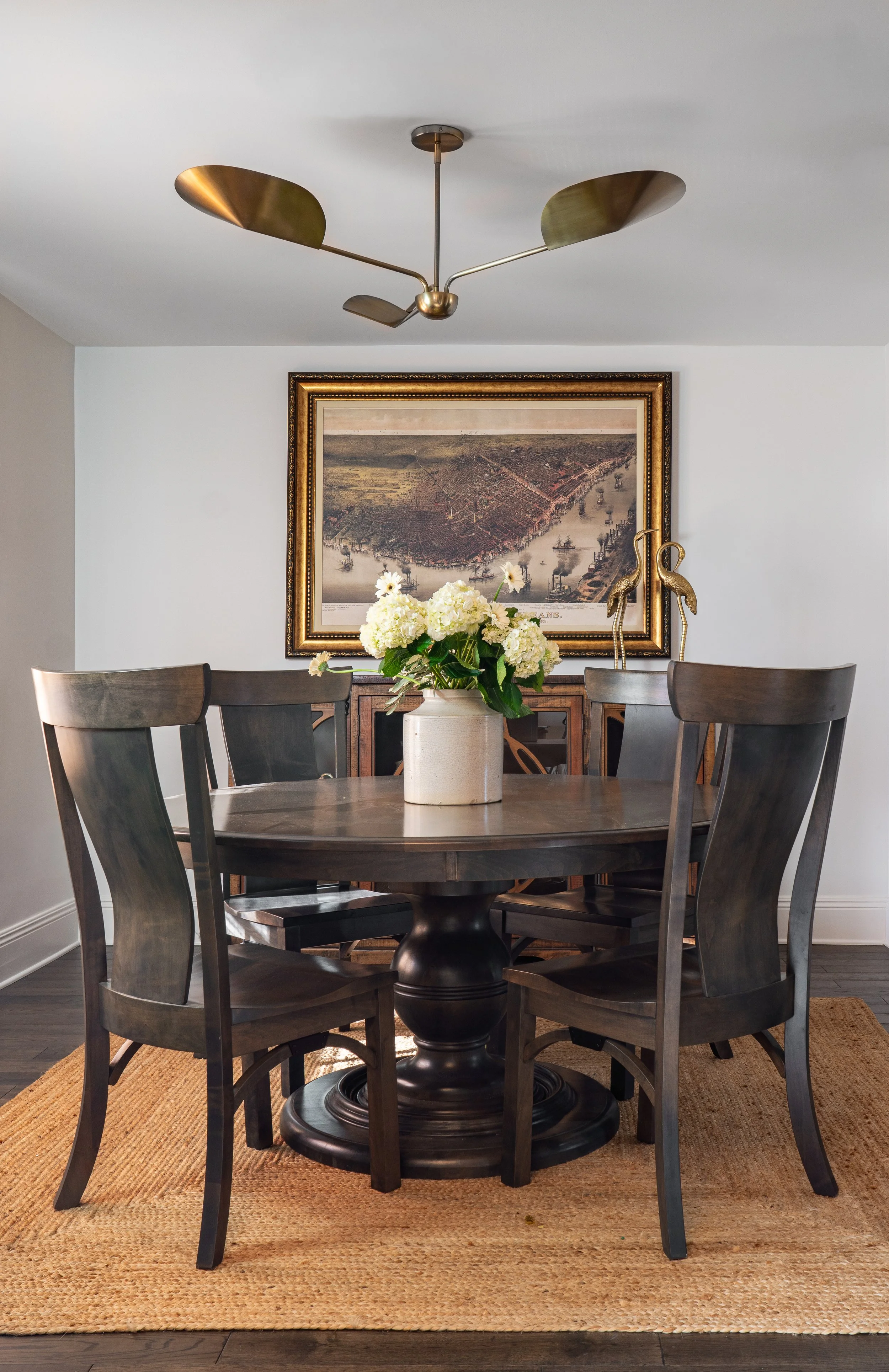 A dining room with a round wooden table, four matching chairs, a white vase with white flowers, a framed map on the wall, decorative brass flamingo figures, and a modern brass ceiling light fixture.