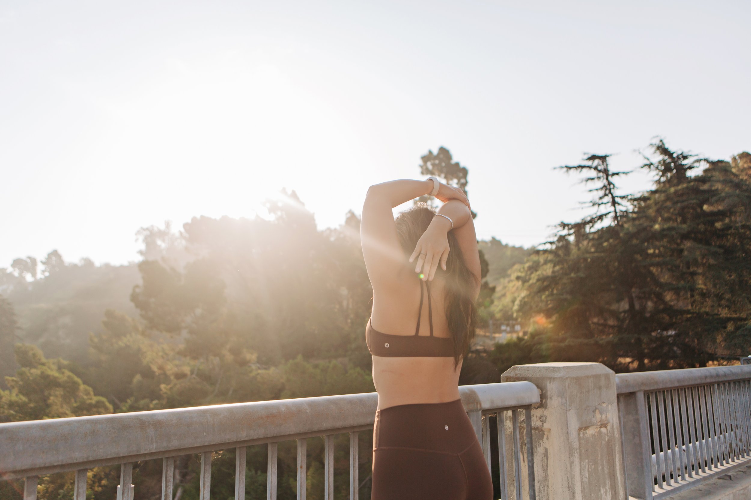 A woman wearing a sports bra and leggings is standing on a bridge with a forested landscape in the background, illuminated by sunlight.