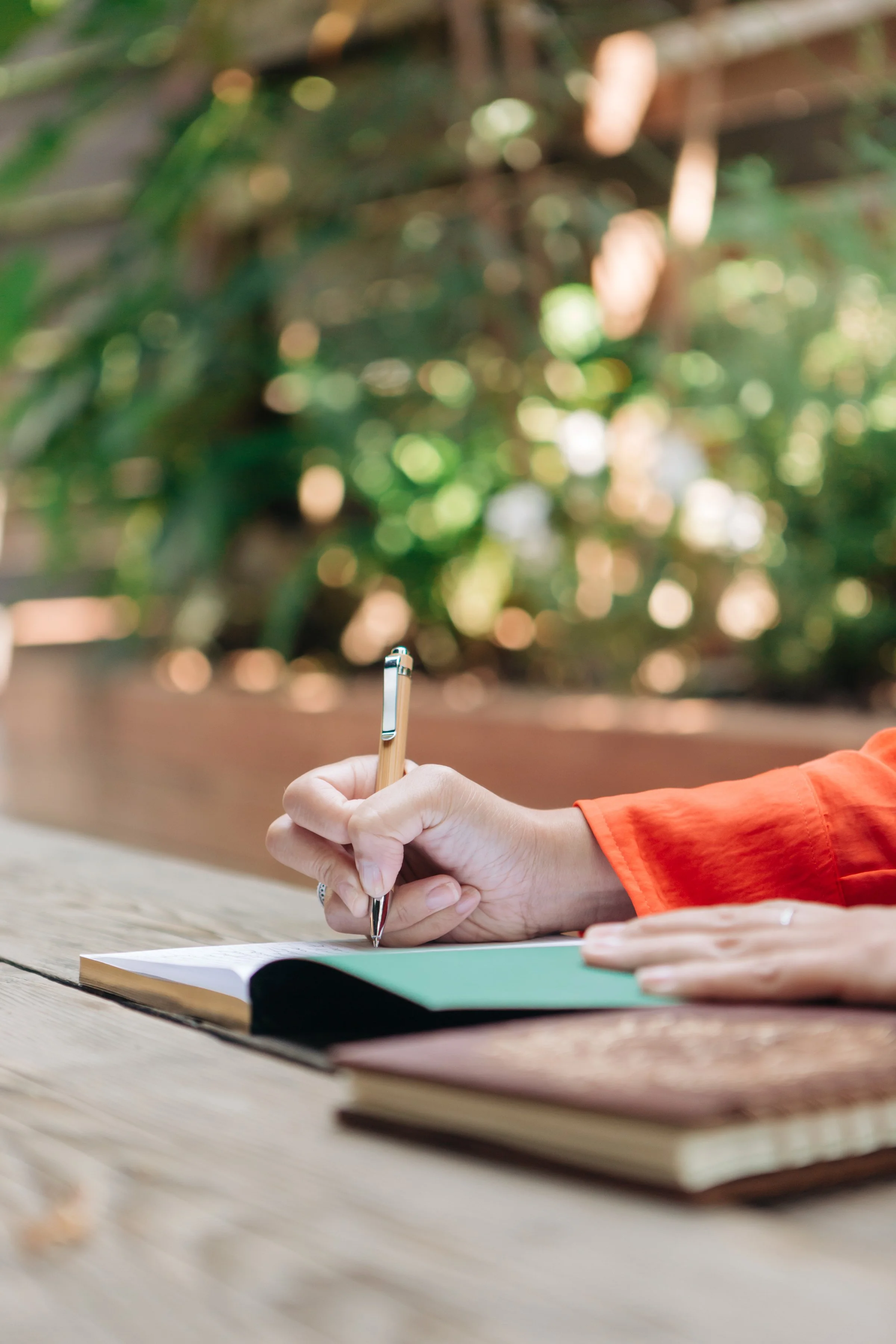 Woman in an orange shirt writing in a notebook at a wooden outdoor table with a small cactus in a pot in the foreground, surrounded by greenery.