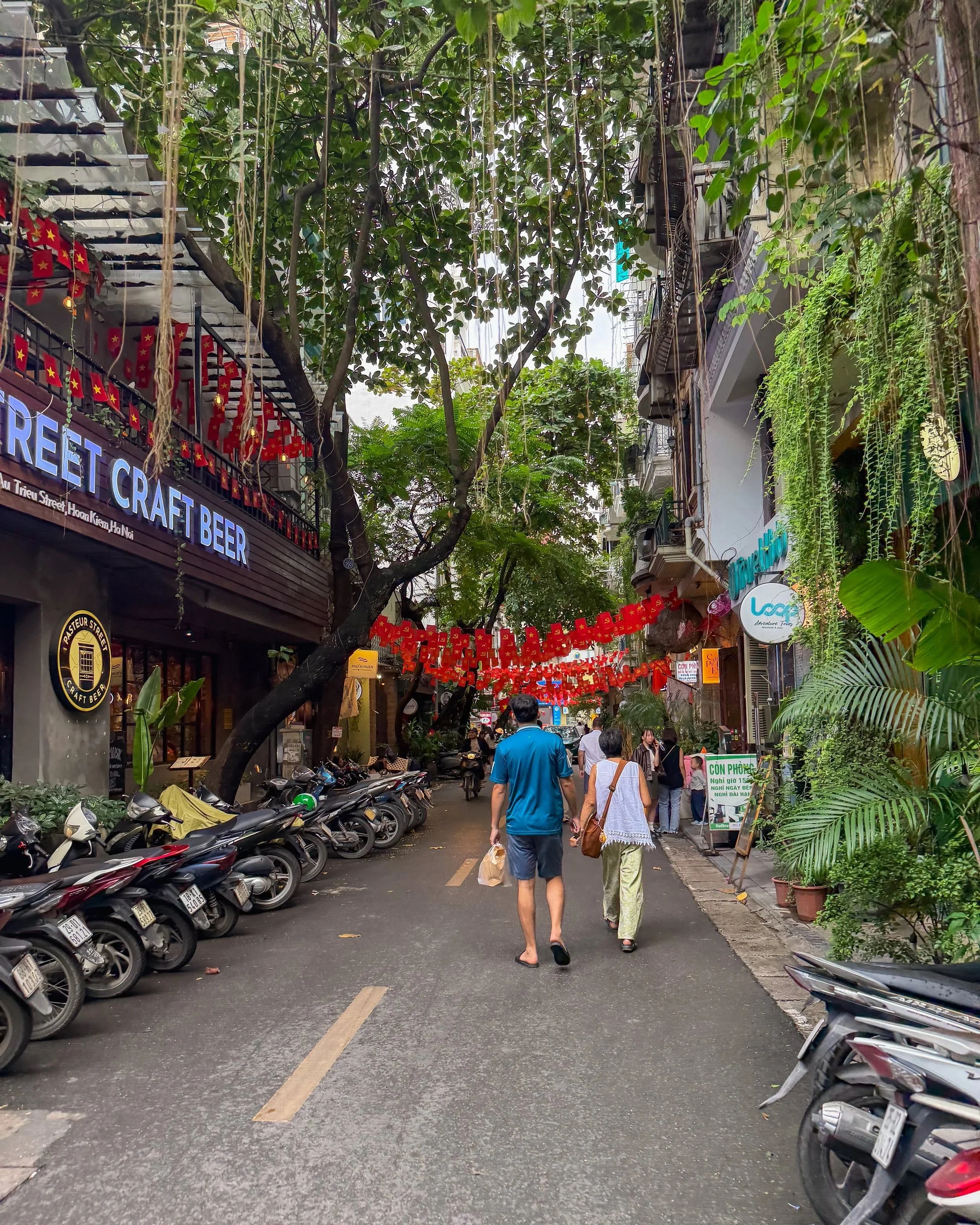A quiet side street in Hanoi lined with lush green plants, parked motorbikes, colorful storefronts, and red banners strung overhead as people walk beneath the trees.
