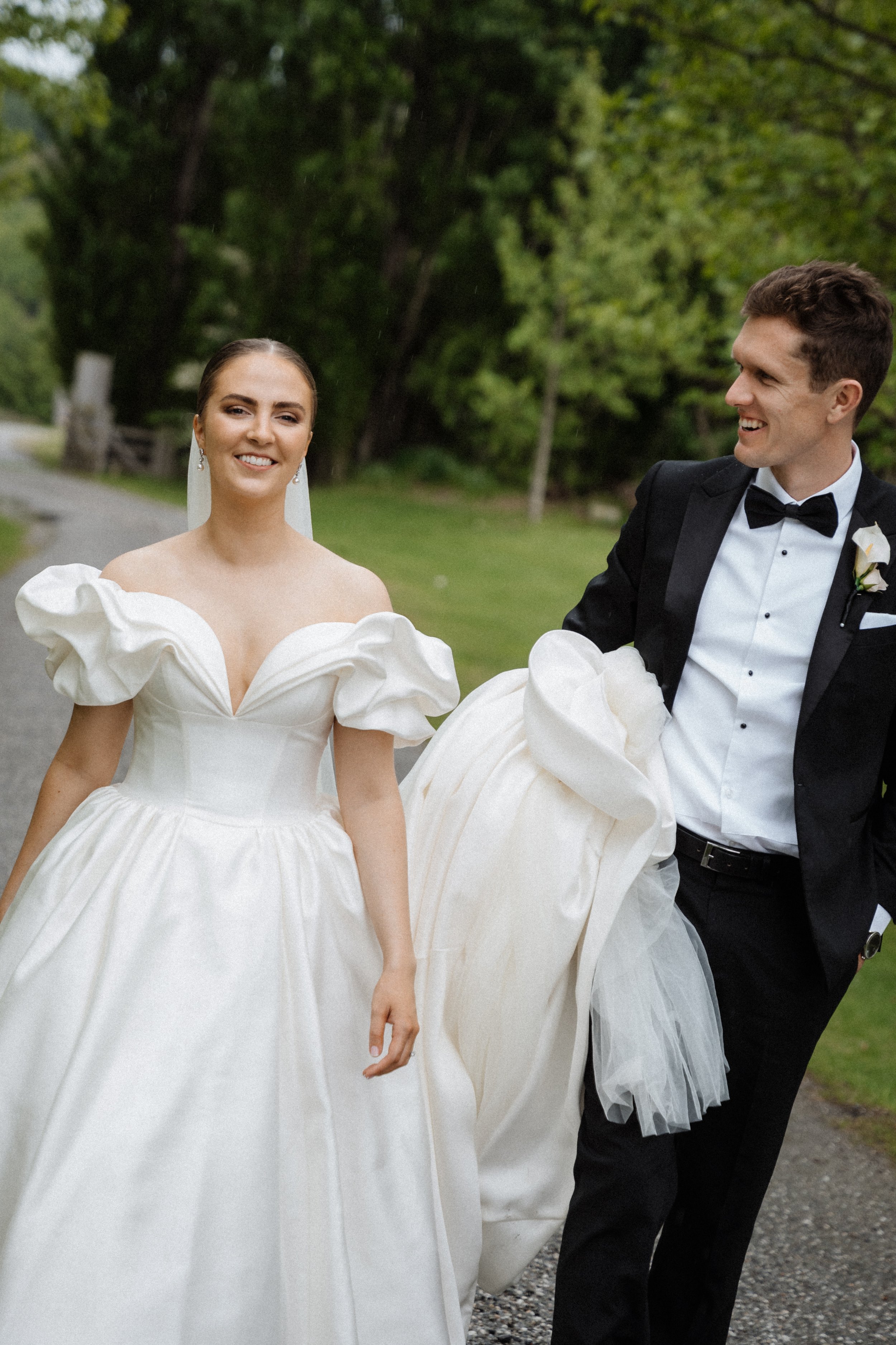 A bride and groom walking outdoors on a gravel path surrounded by green trees. The bride is wearing a white gown with puffed sleeves, and the groom is in a black tuxedo with a bow tie. They are smiling and looking at each other.