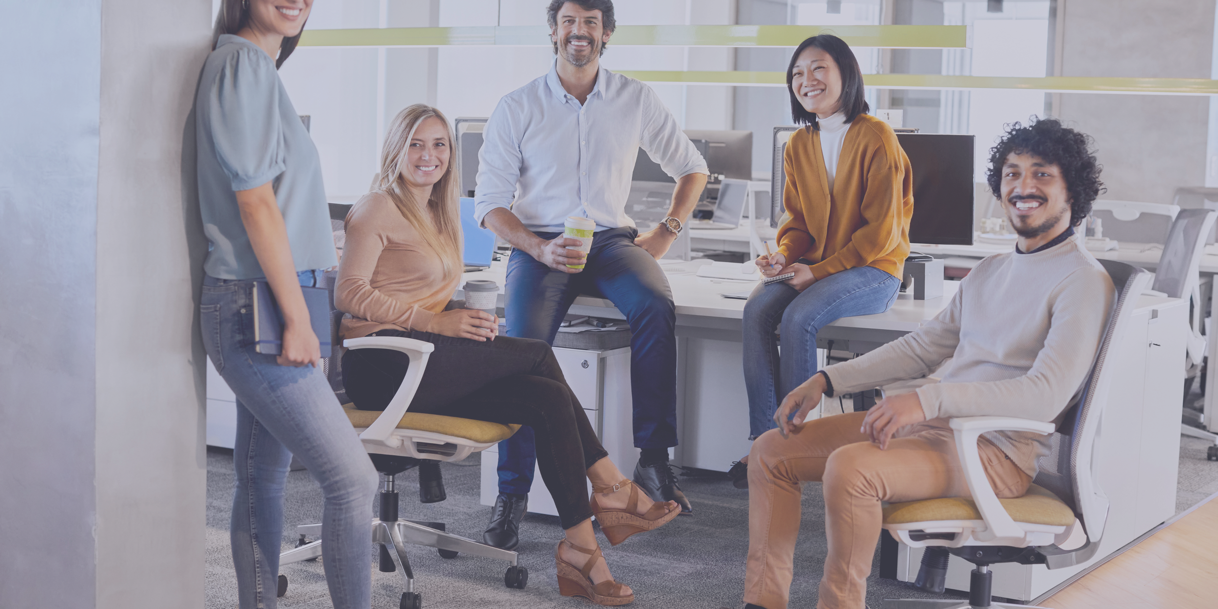 Group of five young professionals smiling and socializing in a modern office with open workspace and computers.