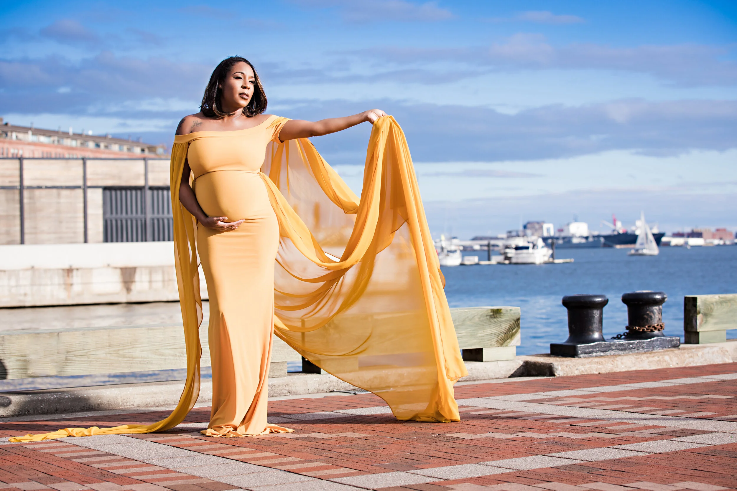 Pregnant woman in yellow gown near waterfront with flowing fabric, boats in background.
