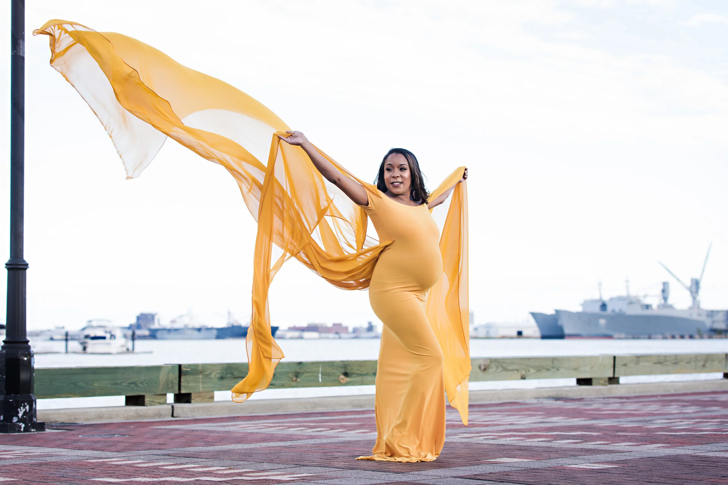 Pregnant woman in yellow dress with flowing fabric posing by waterfront.