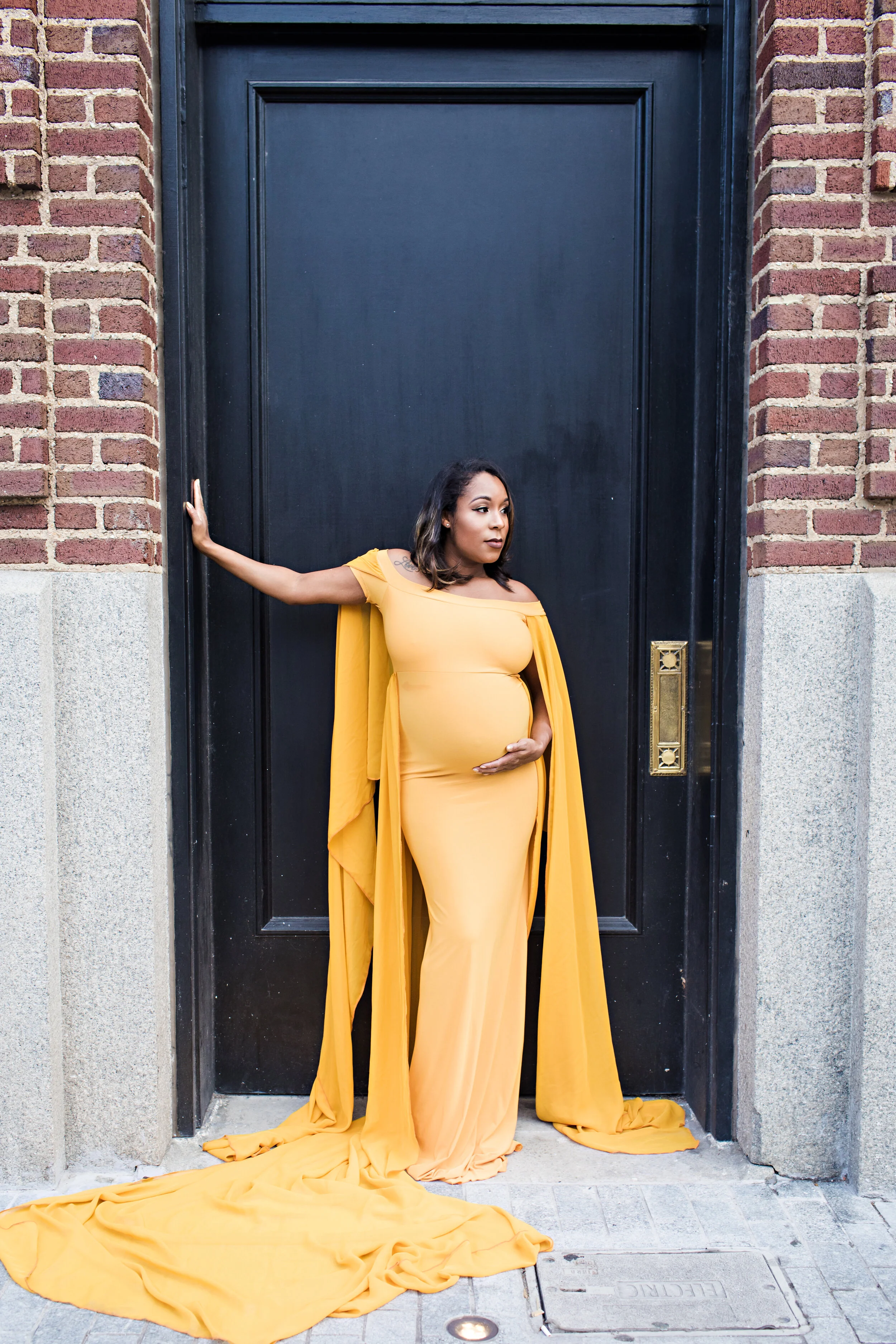 Pregnant woman in a yellow gown posing against a brick wall and door.