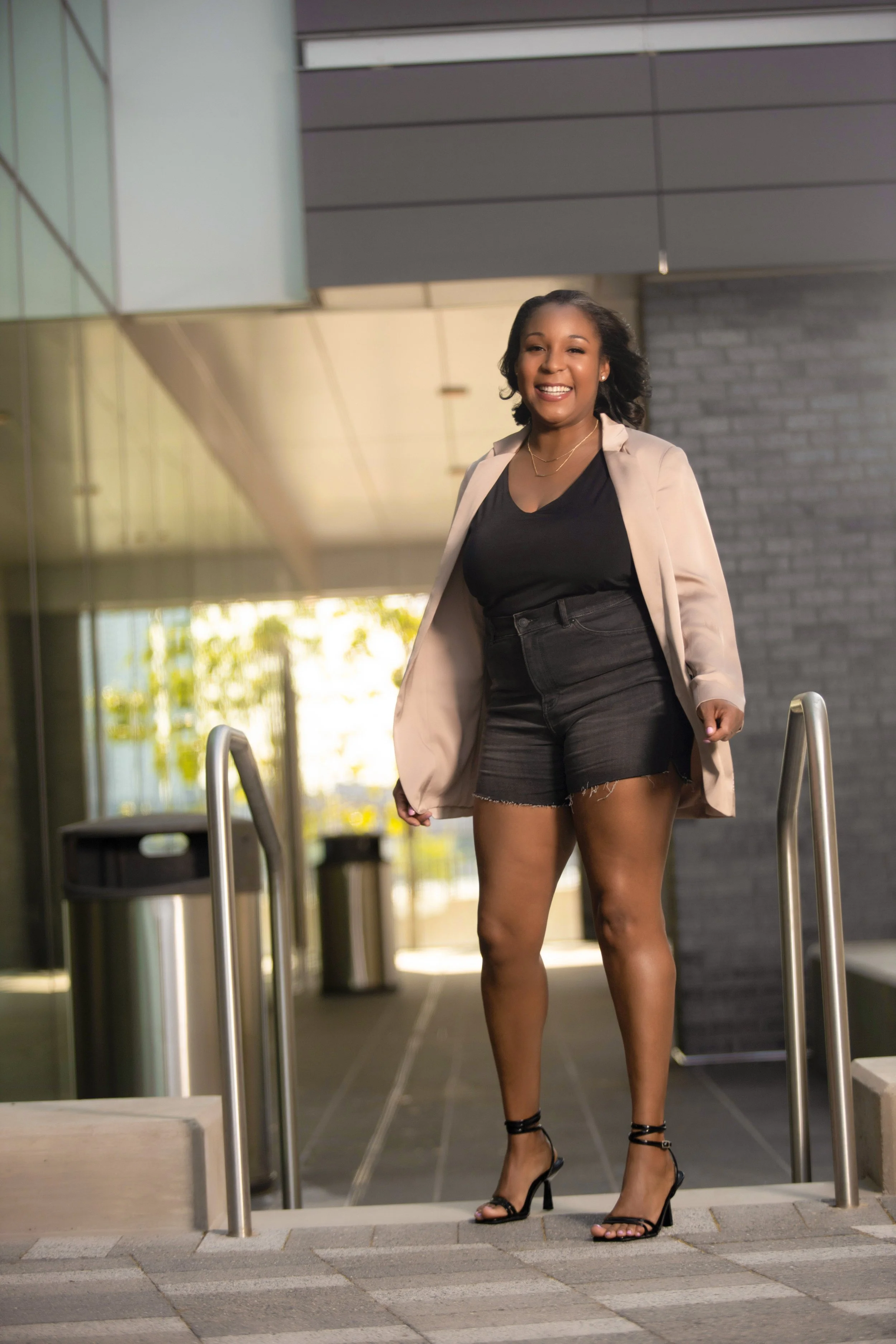 Woman in black top, shorts, and beige blazer standing on steps outside a modern building.