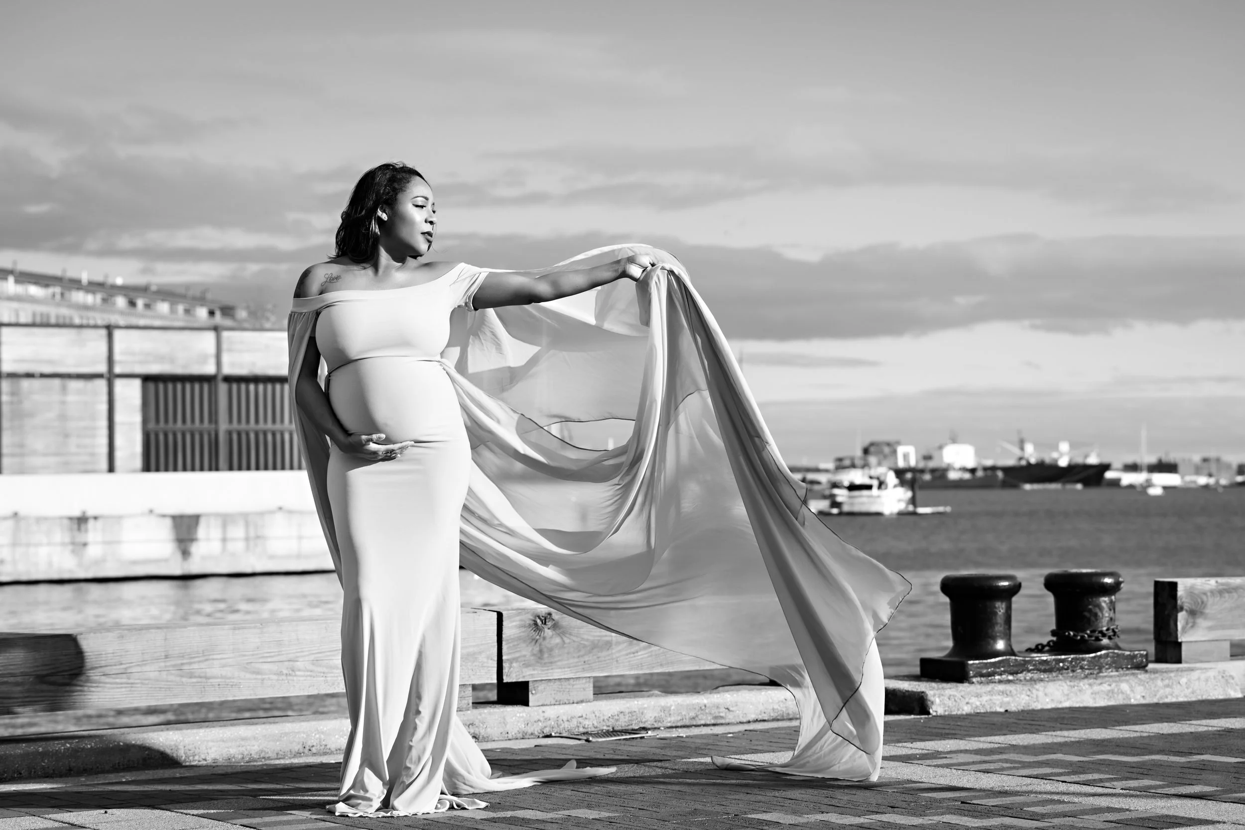 Pregnant woman in long dress at waterfront, holding fabric in wind, black and white photo.