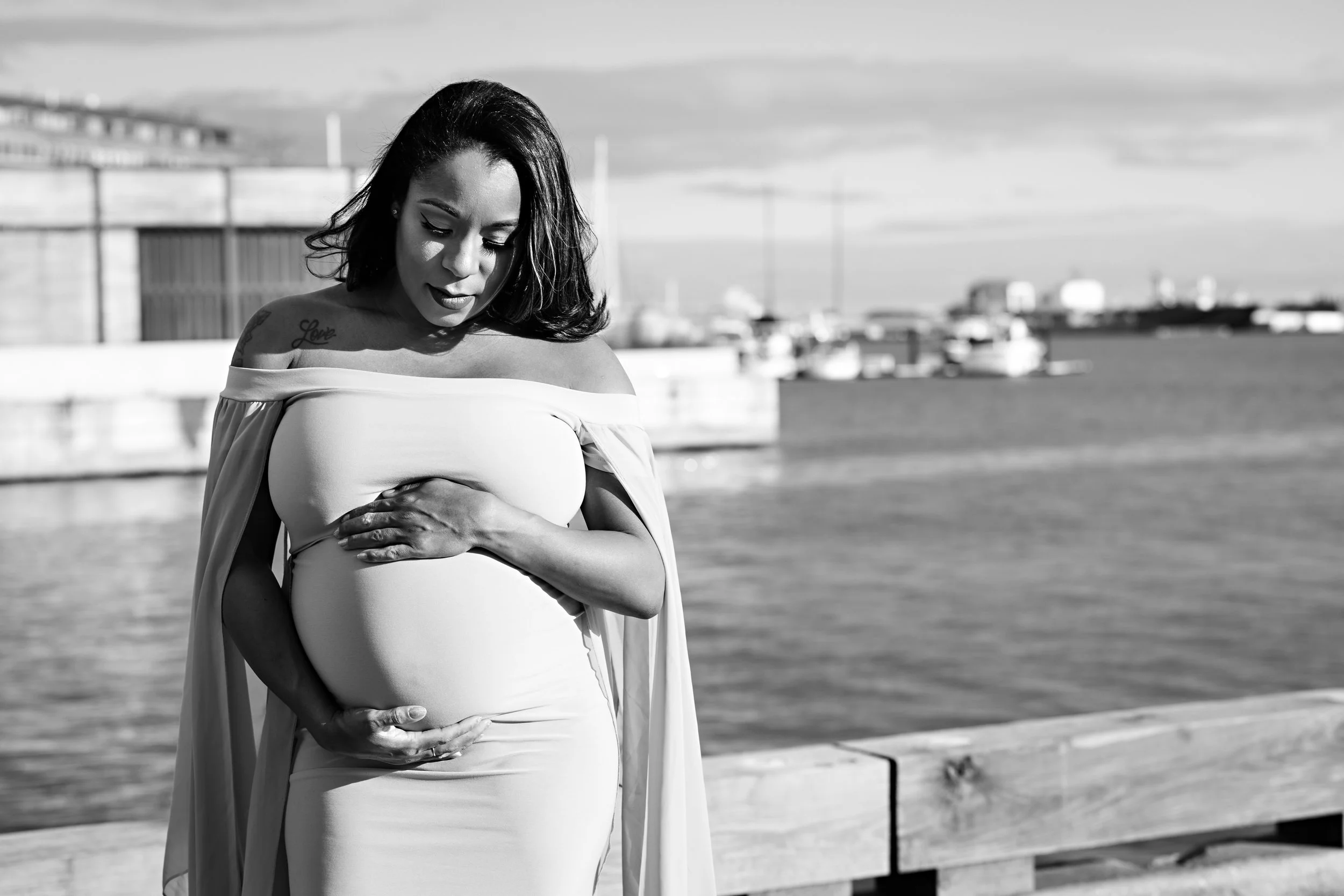 Pregnant woman in off-shoulder dress standing by waterfront, black and white photo