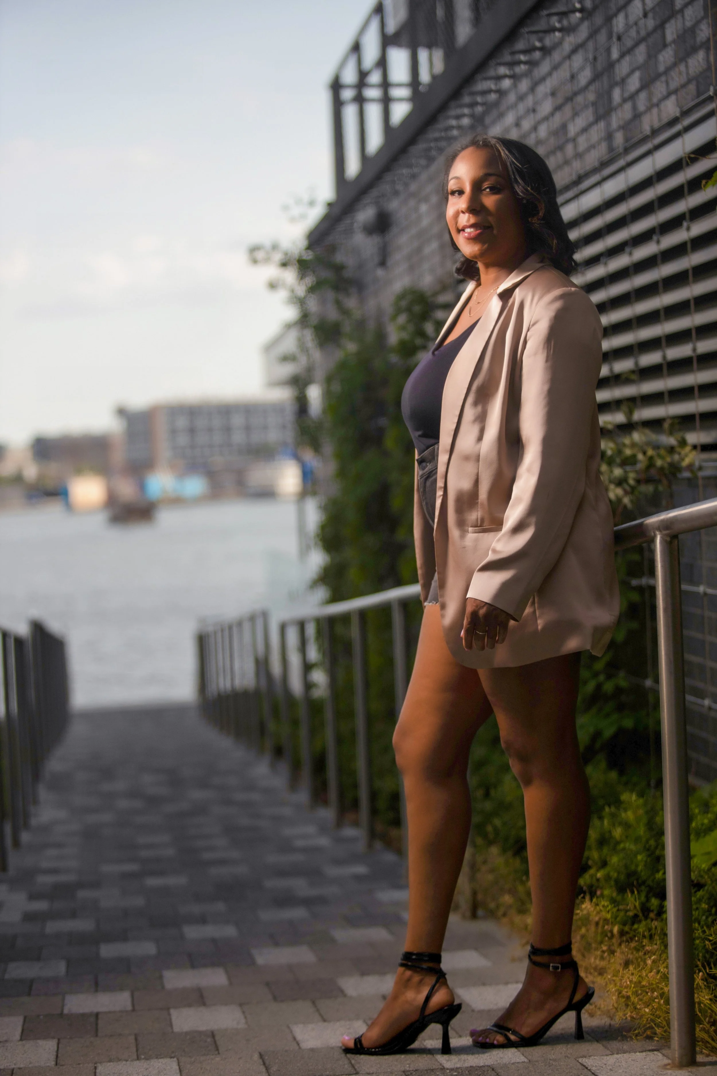 Woman in a blazer standing on a pathway near water, with buildings in the background.