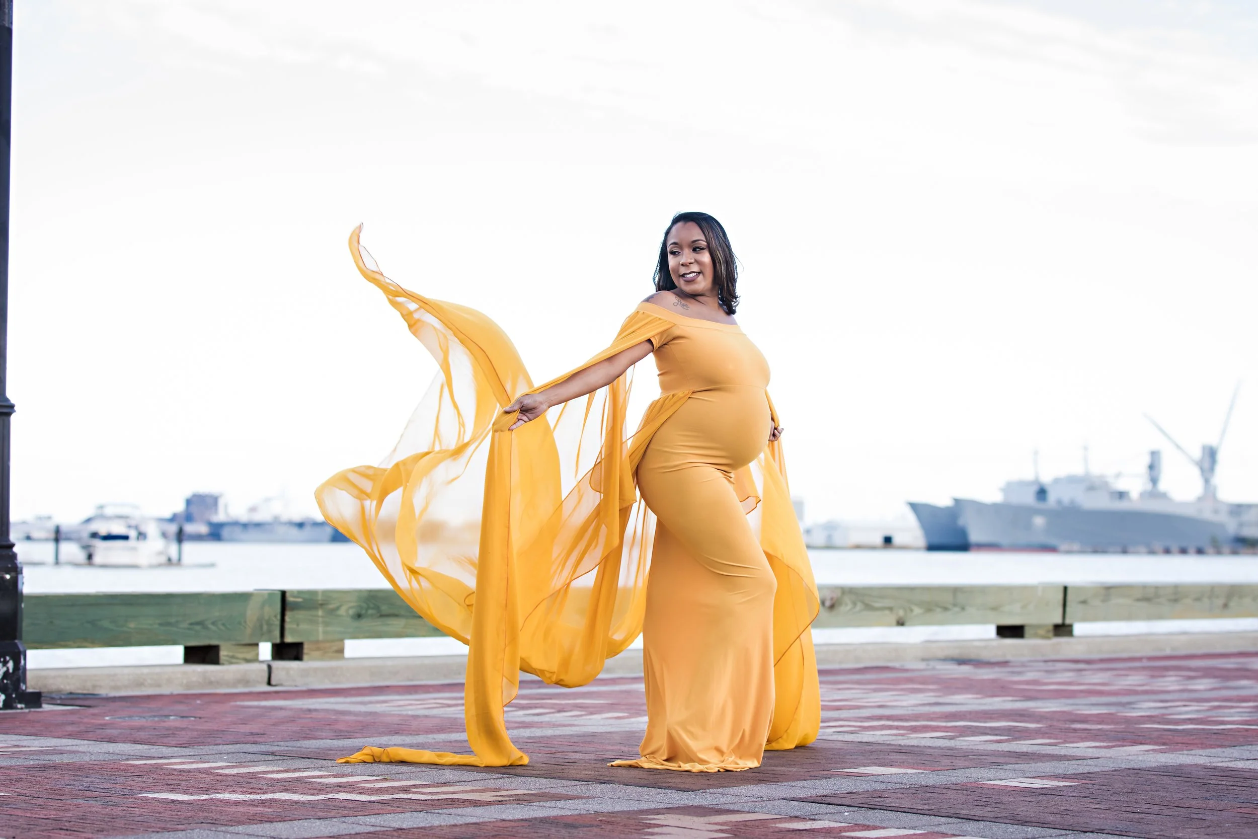 Pregnant woman in flowing yellow dress by waterfront with ships in background.