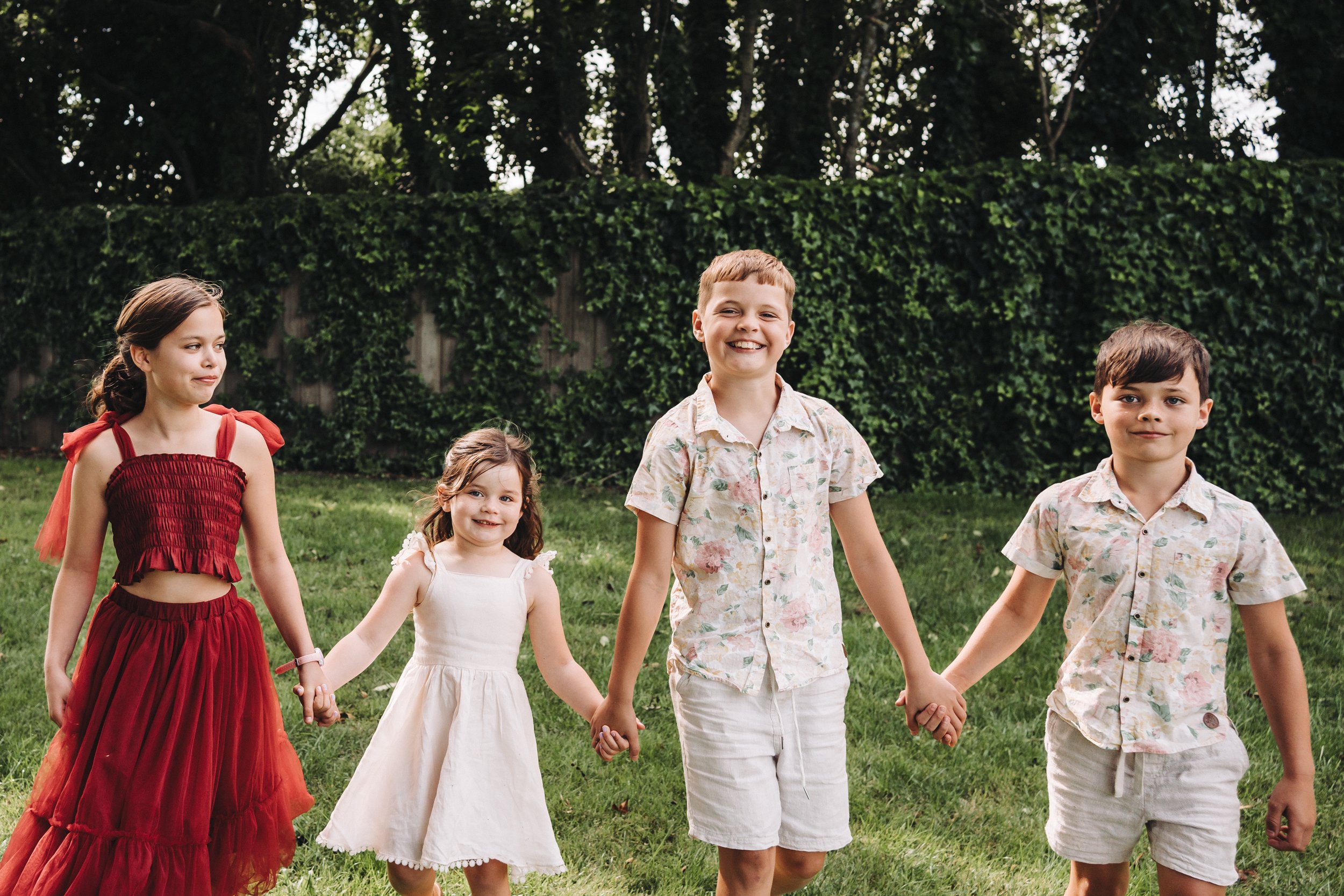 4 sibblings walking hand in hand all looking happy, two girls, one wearing a deep read outfit, the other in a cream dress, the two boys have cream shorts and crea, shirts on, there are trees and posin ivy in the background