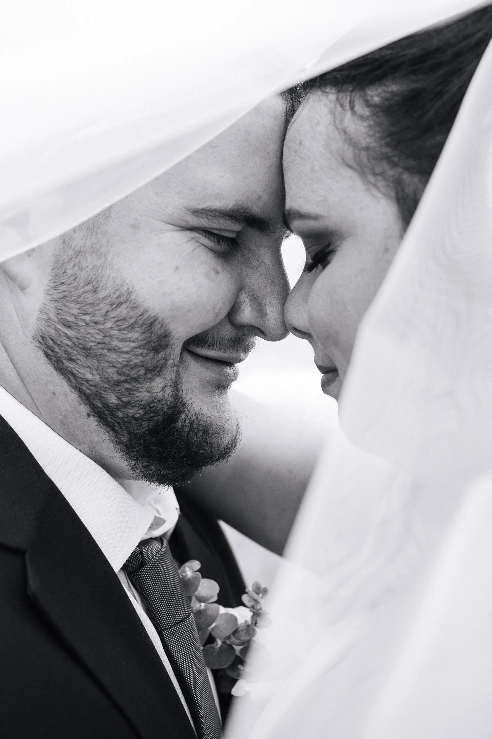 close up of a bride and grooms who are touching foreheads and noses with their eyes closed, smiling, there is some veil fabric framing them and the image is black and white