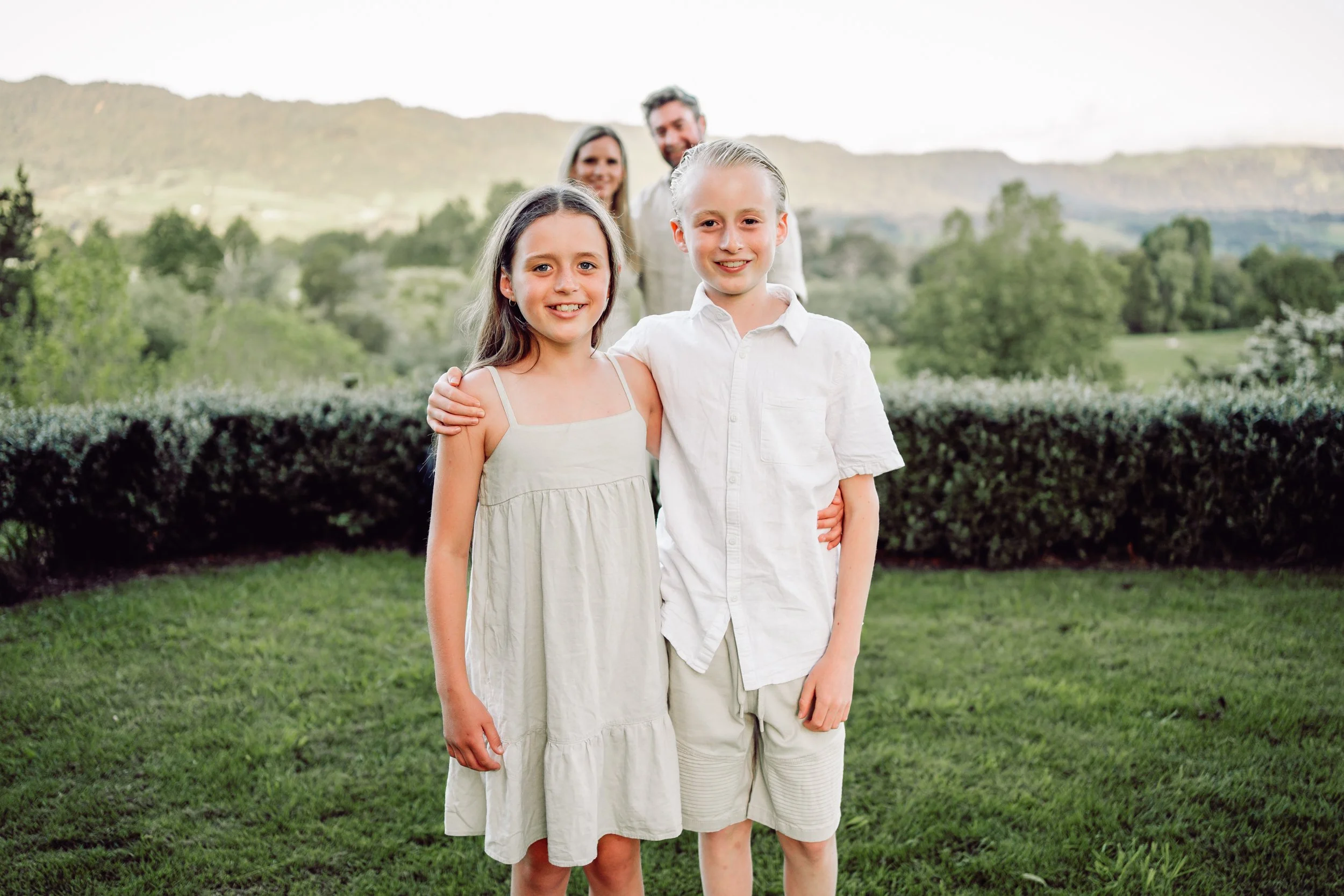 A brother & sister with their arms round each other smiling big smiles standing on a lush green lawn, mum and dad are out for focus in the mid ground and there is the kaimai ranges in the background with the sun light setting pover it