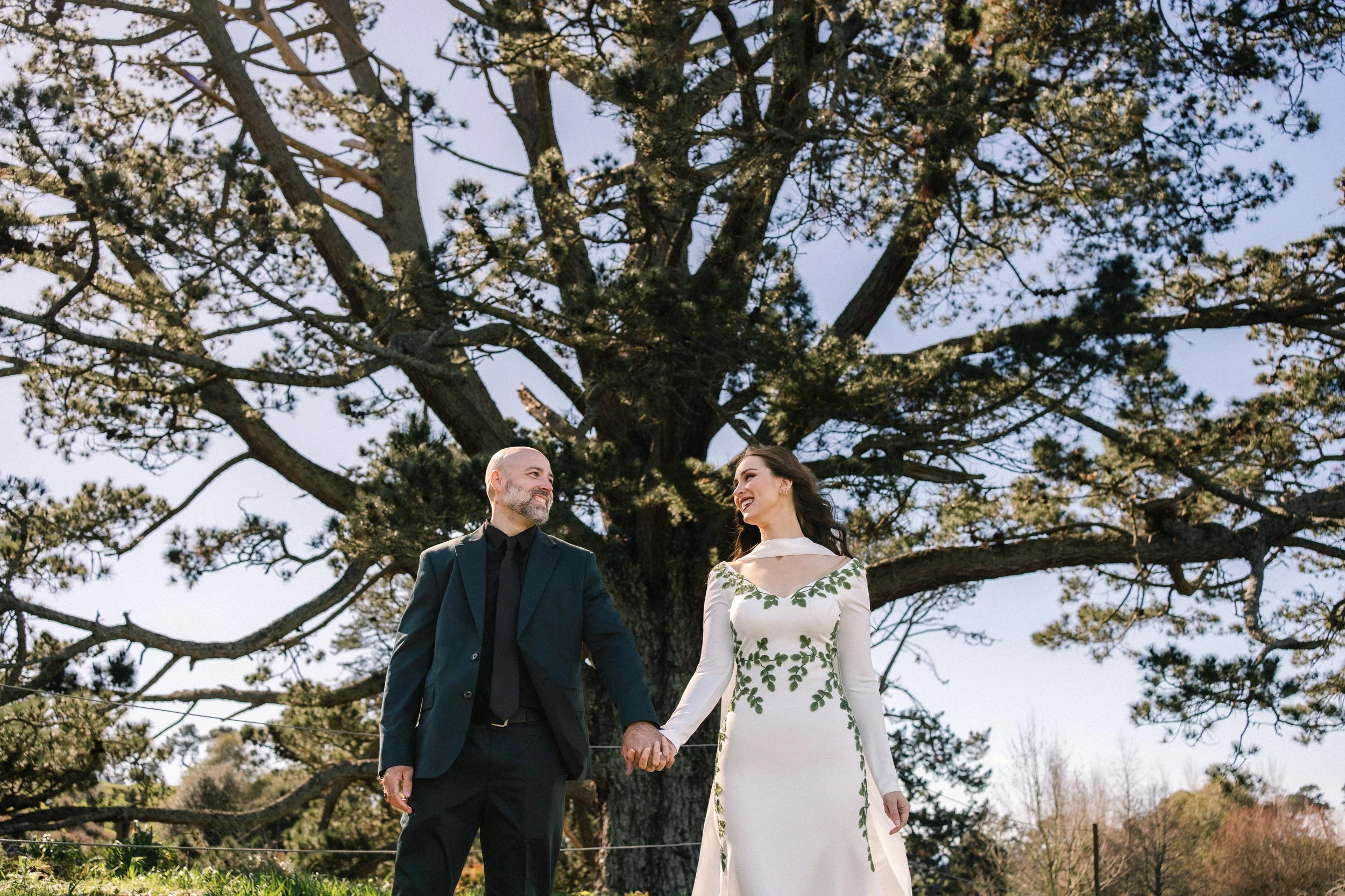 a bride, wearing a long white dress, with green vine detailing and a groom in a navy blue suit, standing in front of a large tree, they are side by side, holding handing hands, looking at each other and smiling with their teeth