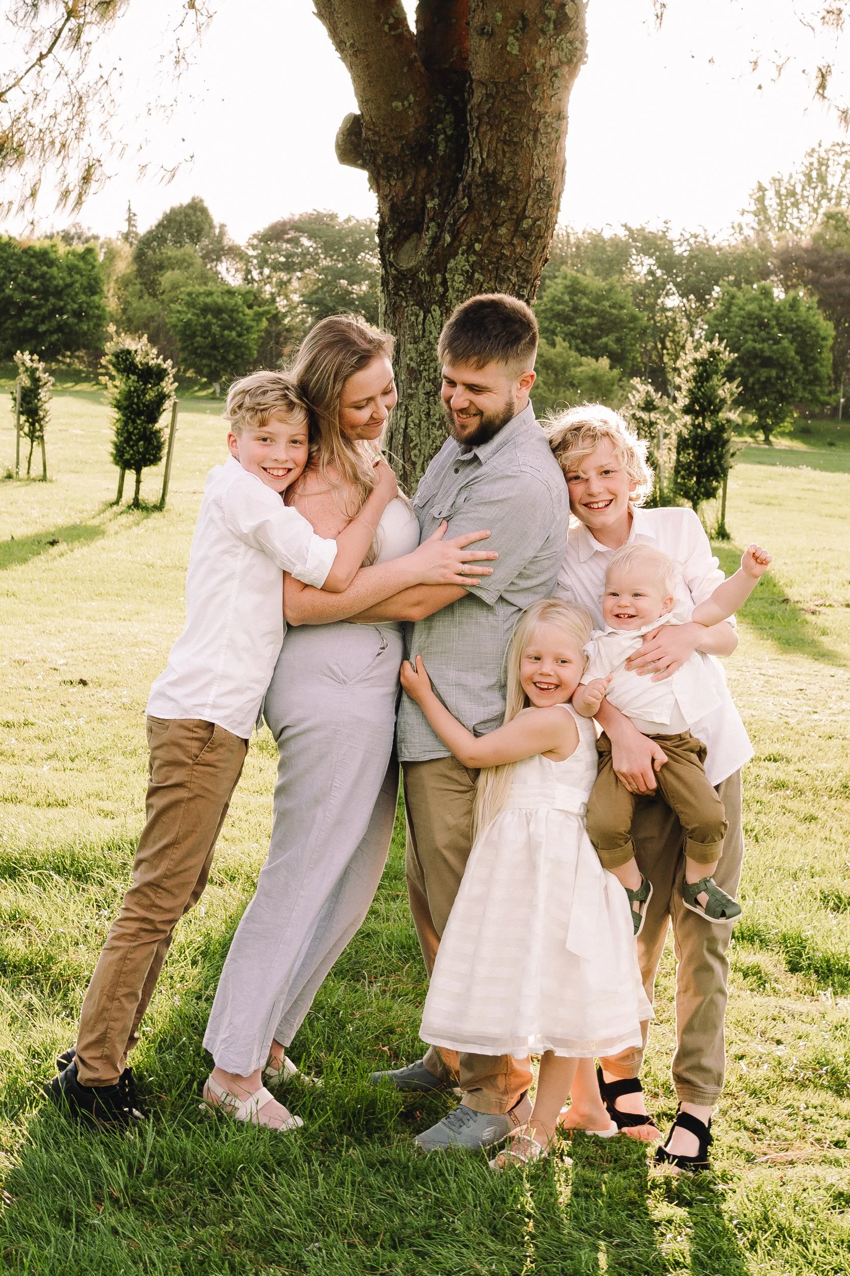 family portrait, mum and dad in the middle chest to chest under the setting sun and in front of a large tree, the 4 children are are ound the outside of them snuggling in and smiling, like a group hug, taken in Matamata, Waikato