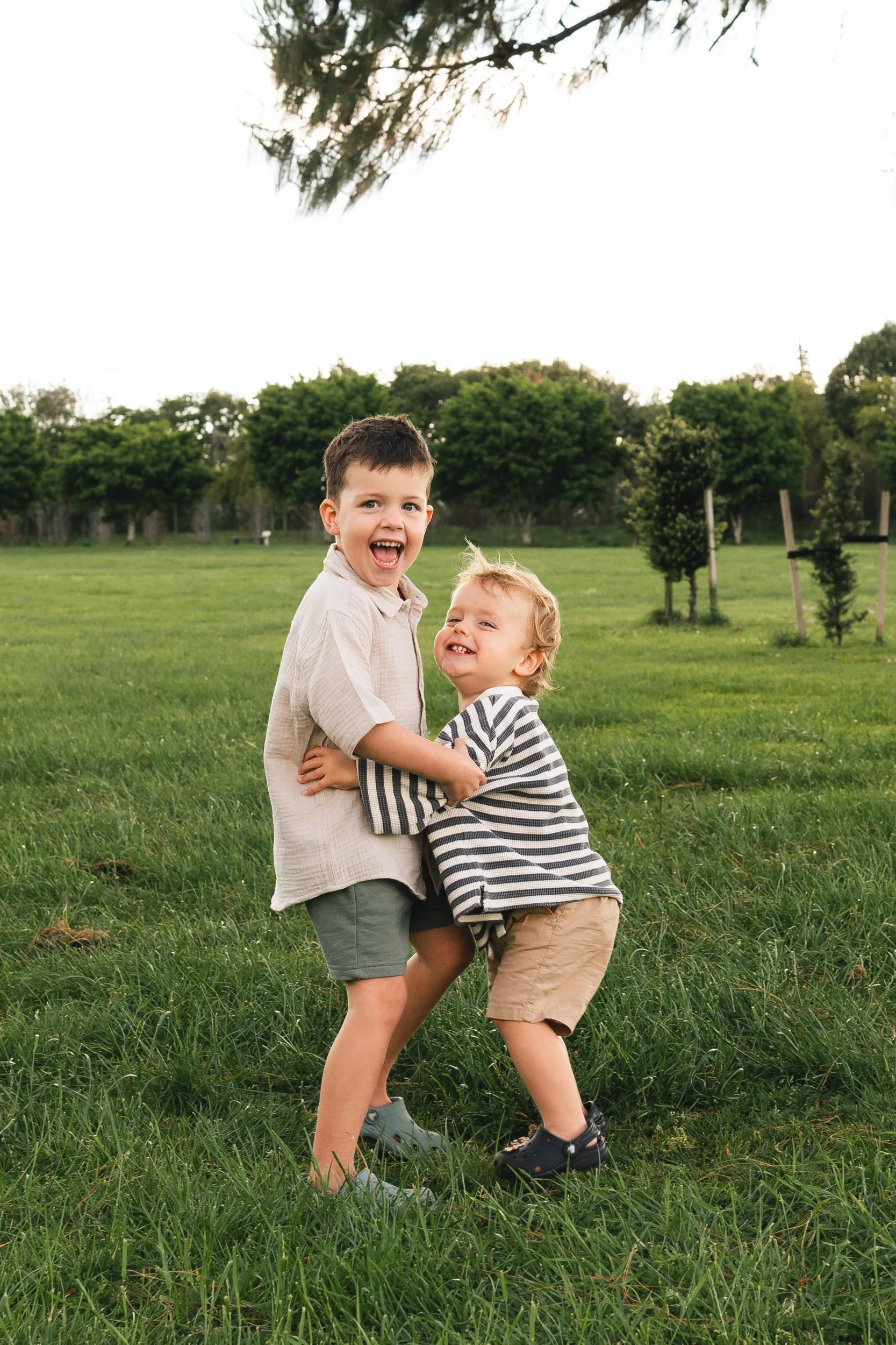 Two young brothers holding each other and laughing, having fun in a lush green field with trees in the background