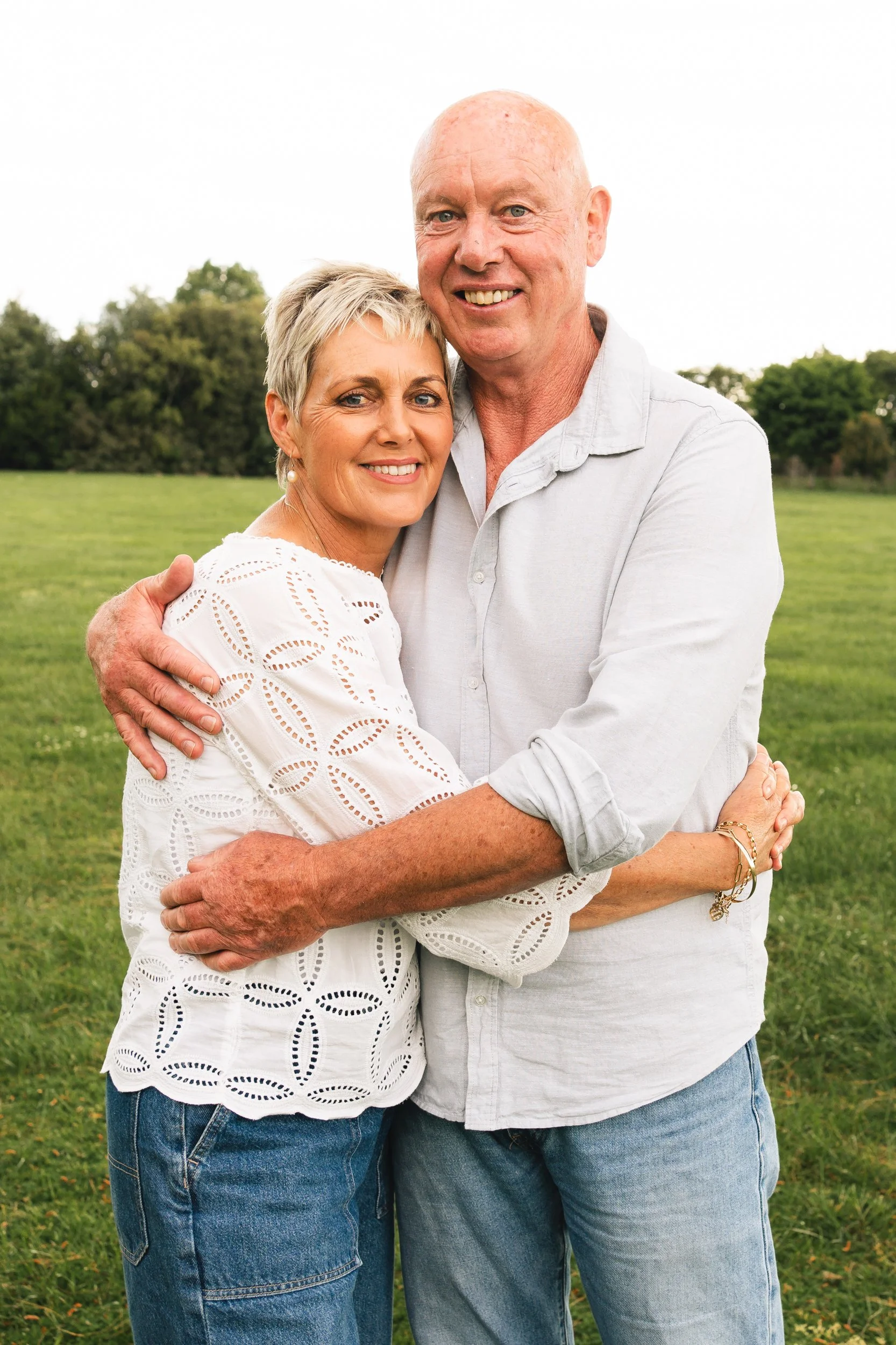 A couple both wearing creamy shirts and jeans, have thier arms wrapped around each other smiling and looking at the camera. Photo taken in Matamata