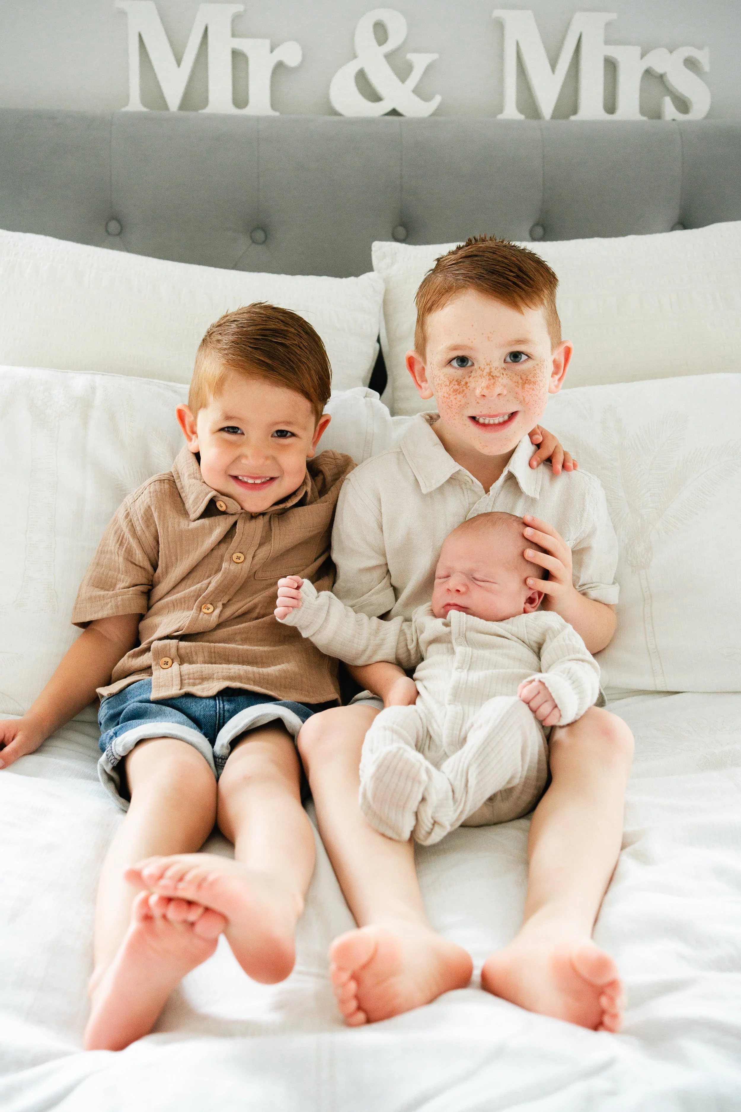 Two little boys sitting on a bed looking at the camera smiling, one is holding his newborn baby brother, they are on a white blanket leaning against white pillows