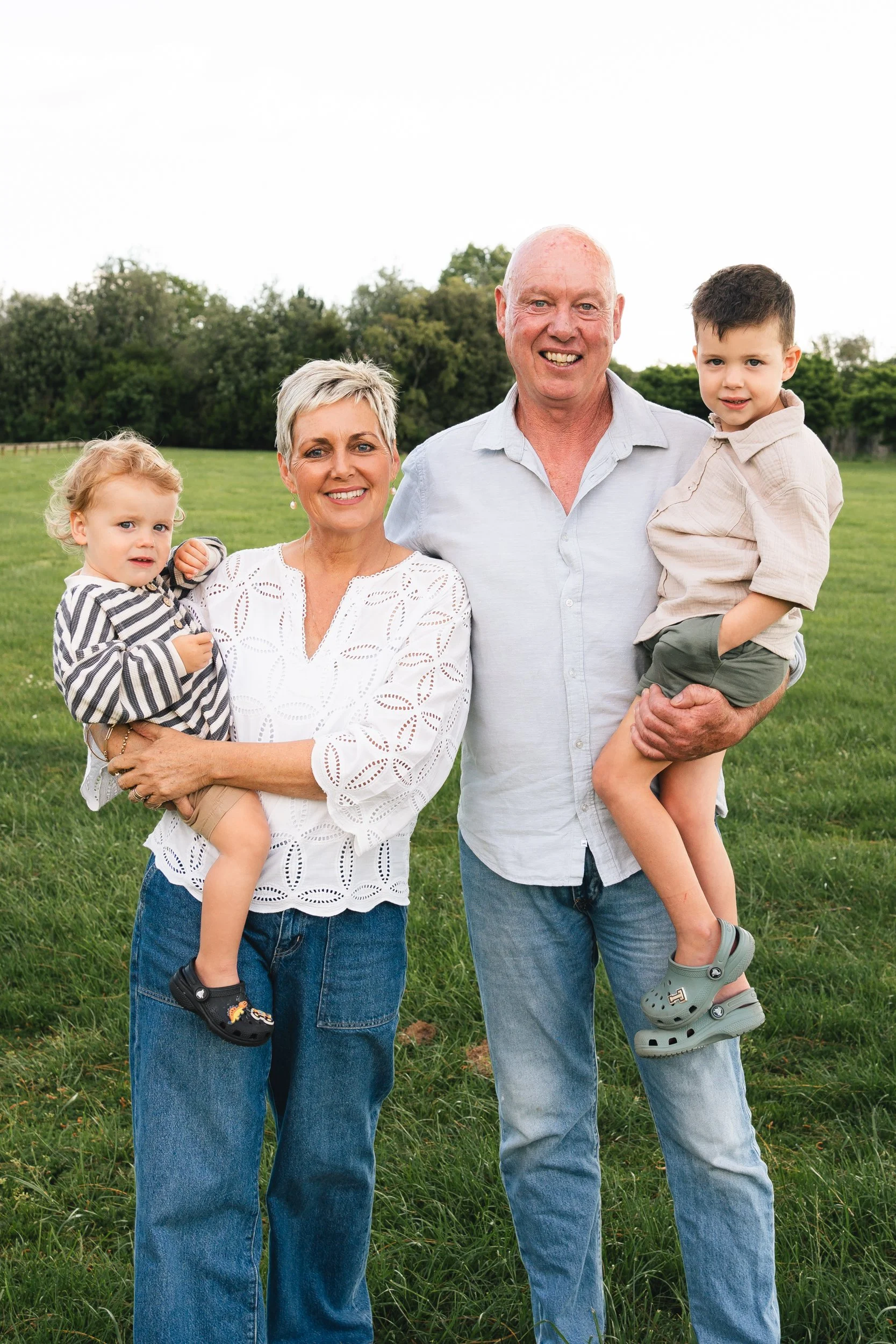 Grandparents standing in the middle, each holding a grandfchild, two boys on their hips, they are standing on lush green grass with trees in the background, photo taken in Matamata, Waikato