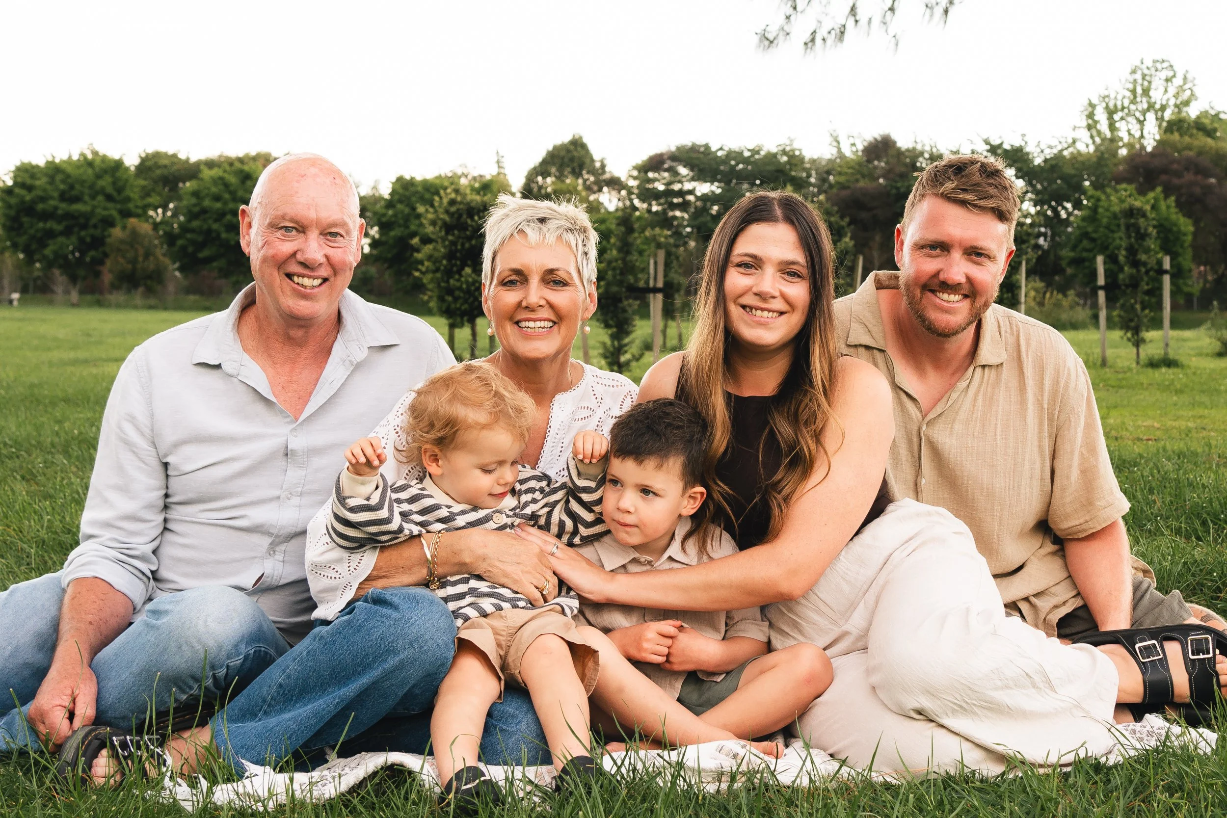 family portrait mum and dad, 2 kids and two grandparents sitting down on the grass with trees behind them the adults smiling and looking at the camera and the kids looking at each other taken in Matamata
