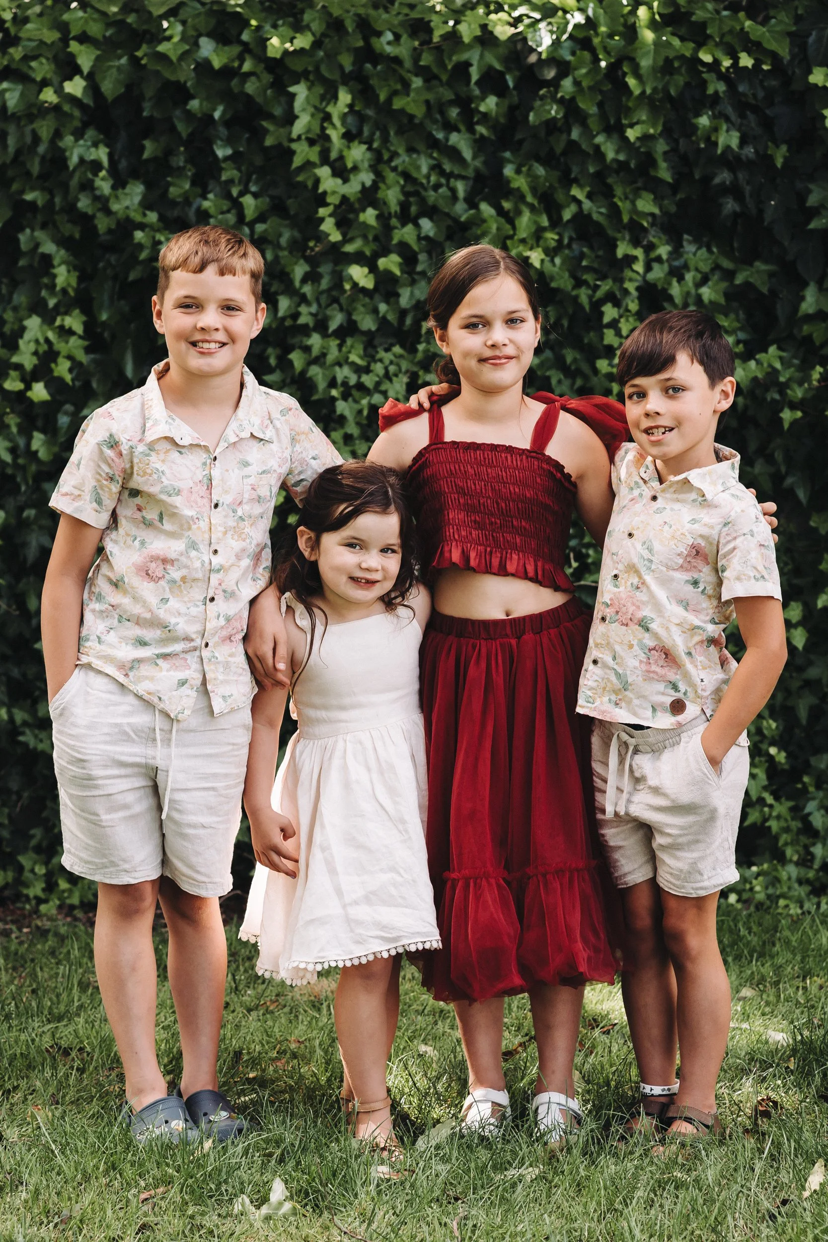 Four siblings standing with their arms around each other, all smiling looking at the camera, on wears a deep red outfit and the others are in cream colours, two brothers and two sisters. Photo taken in Matamata, Waikato