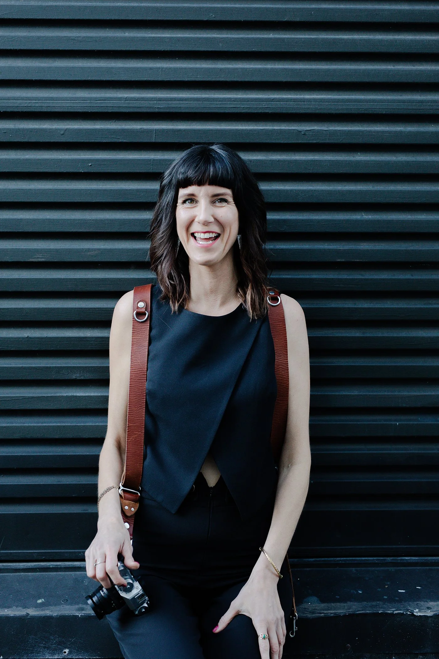 portrait of the photographer, sacha capeling, she is standing in front of a industrial dark metal door, she is smiling a big smile, has black clothes, brown leather camera straps over each shoulder, black hair with bangs