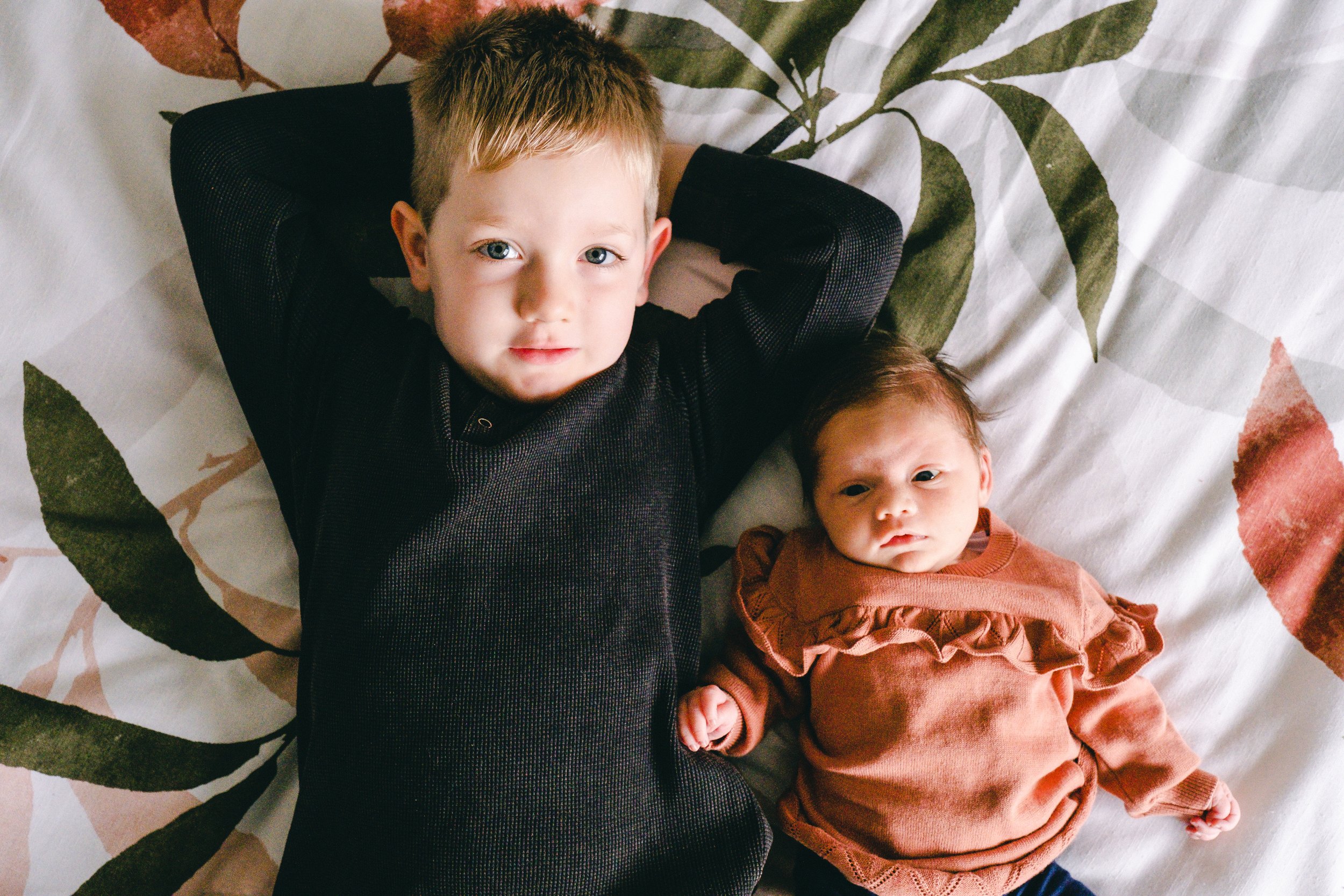 big brother with his arms behind his head and little sister laying on their back looking into the camera.