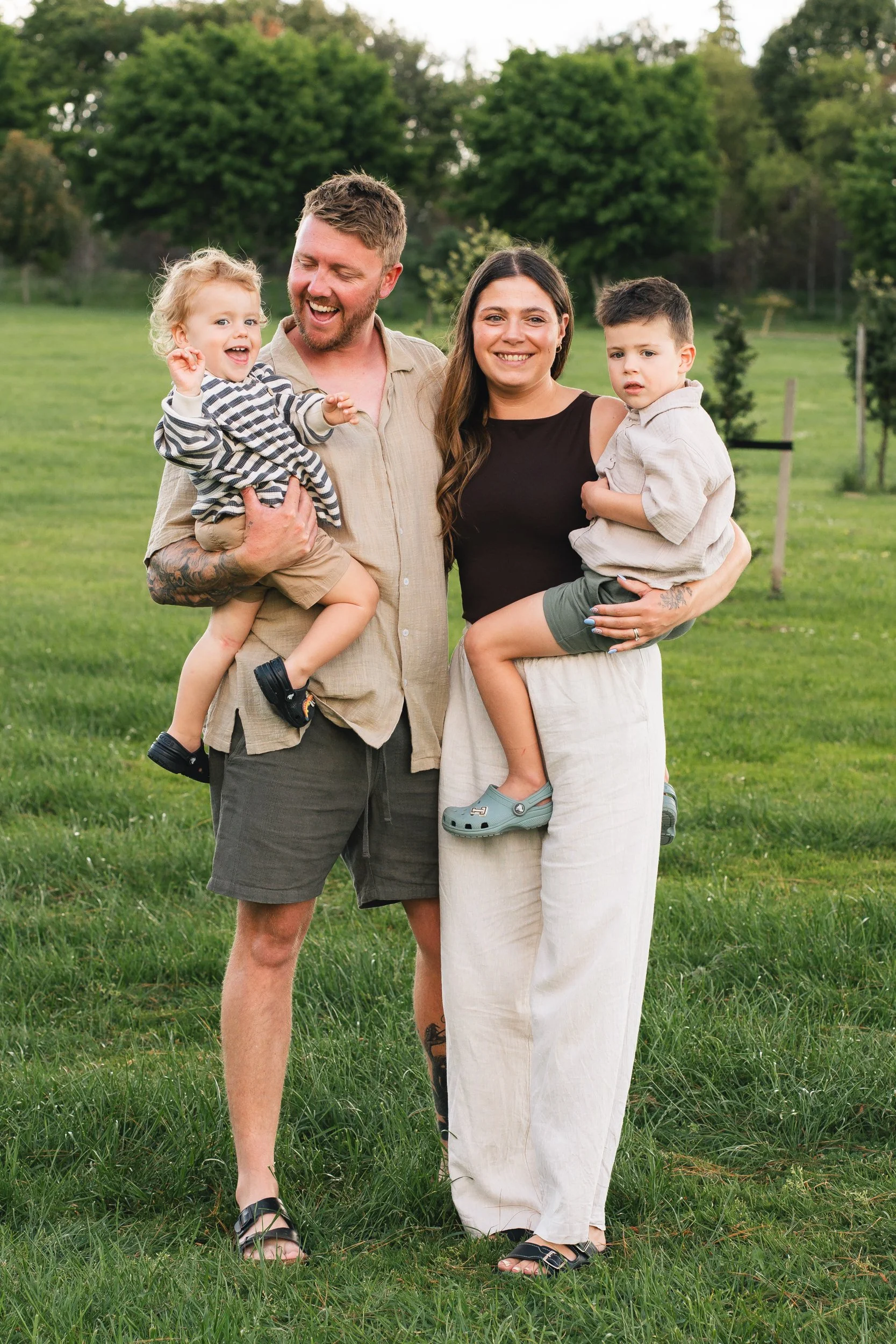 A family of four, mum and dad standing in the middle, each holding a child, two boys on their hips, they are standing on lush green grass with trees in the background, photo taken in Matamata, Waikato