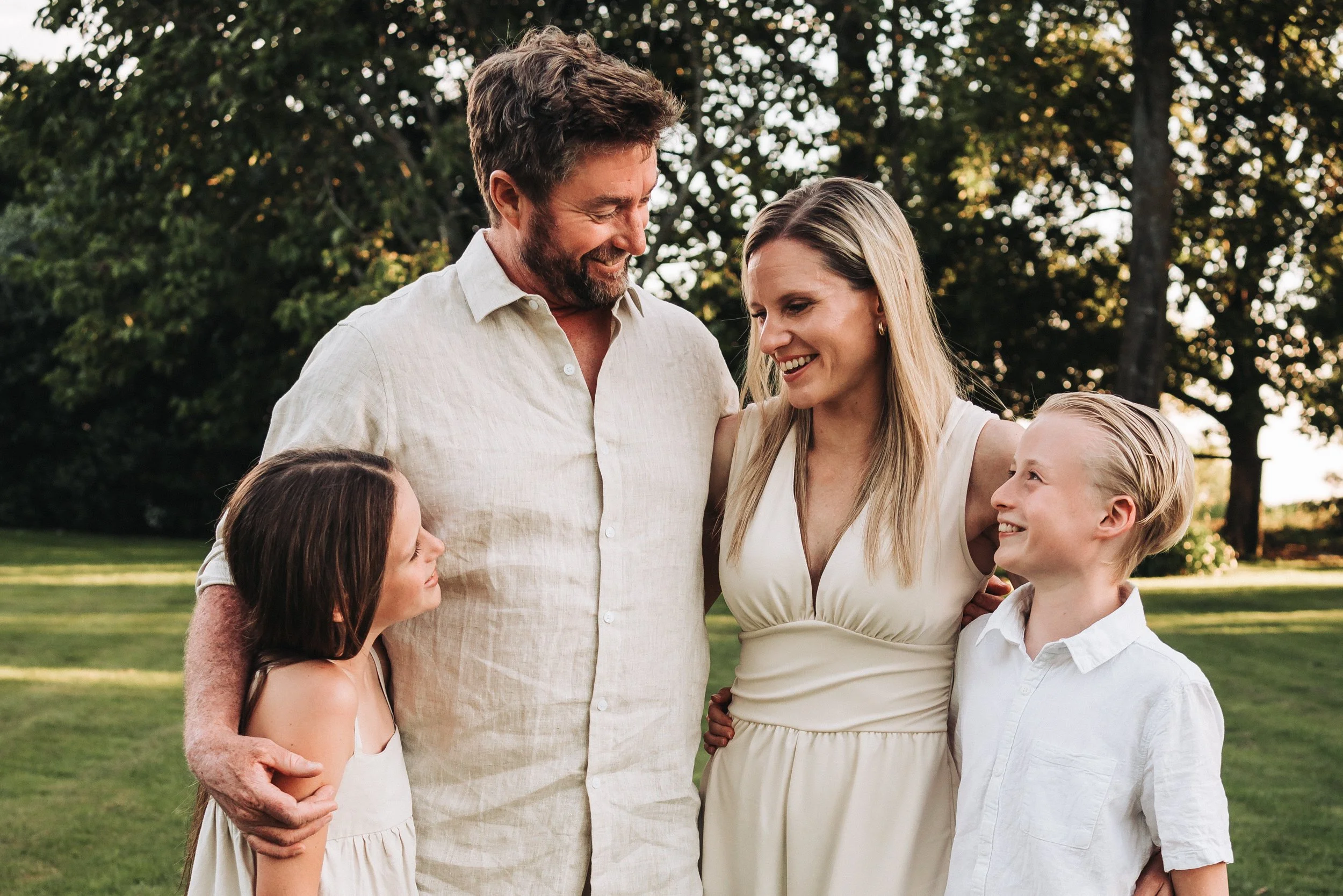family of 4 with their arms around each other, mum and dad in the middle and the daughter and son on either end, all looking at eachother and smiling, large trees in the background