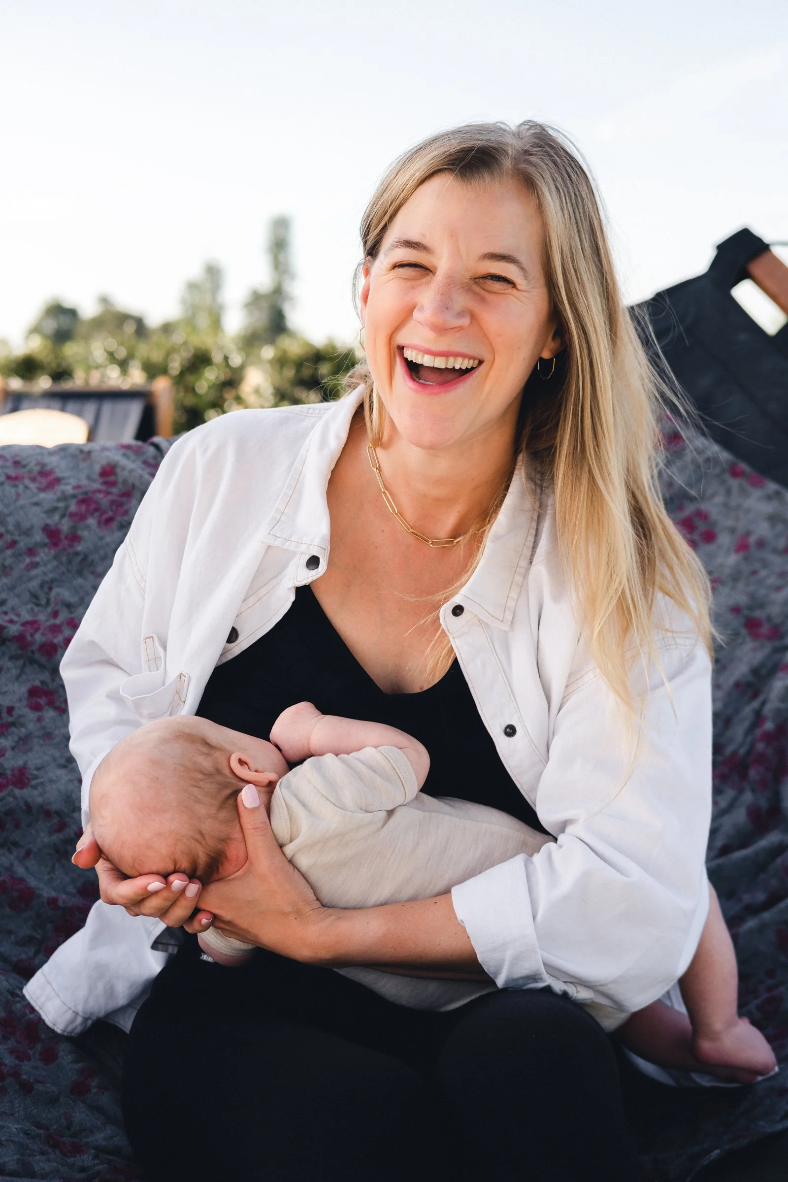 smiling blonde women in white shirt with black trousers breastfeeding her newborn baby