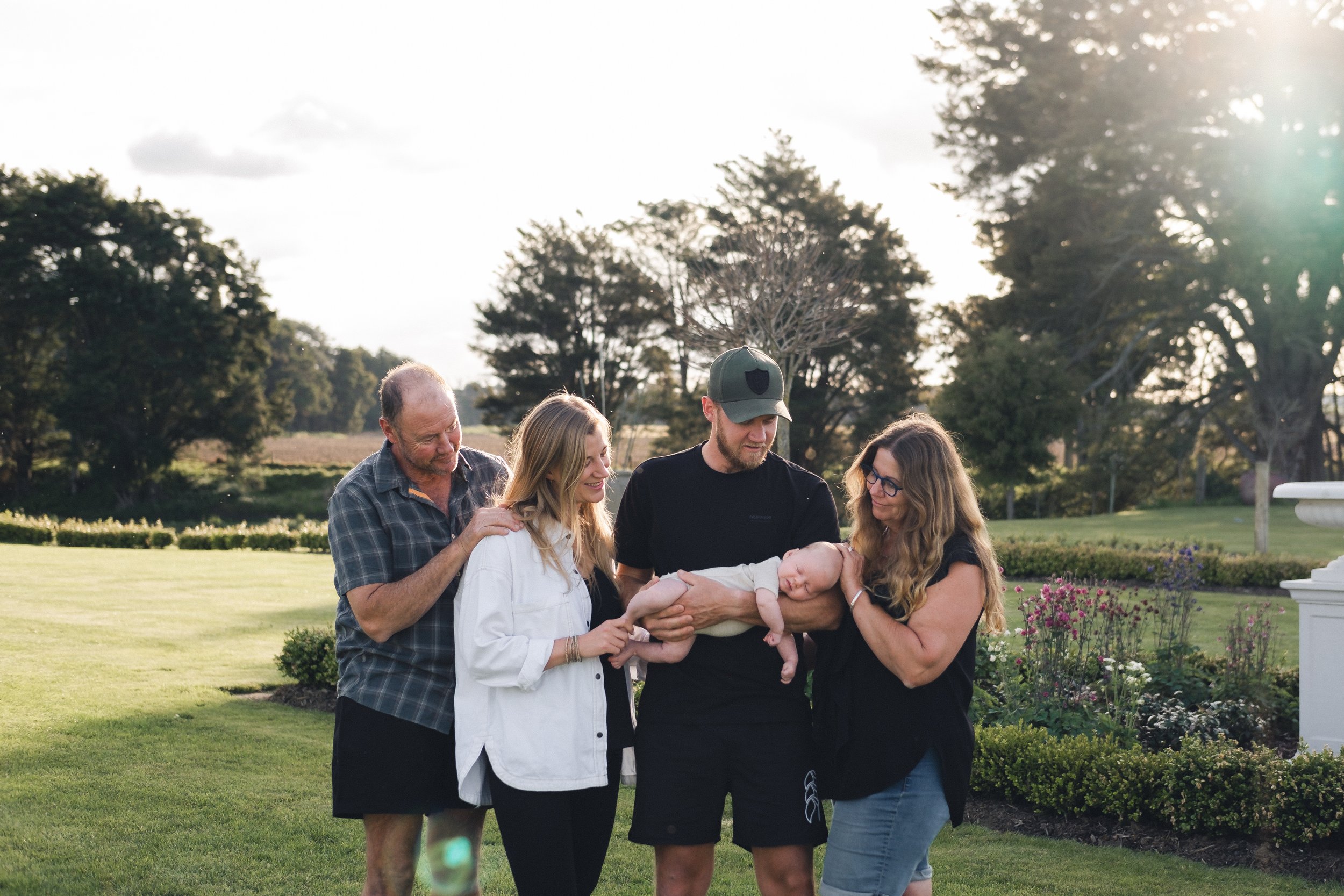 a family photo with two parents and their newborn baby i the middle and the grandparents on the outside. they are all looking at the baby and smiling surrounded by green grass and trees