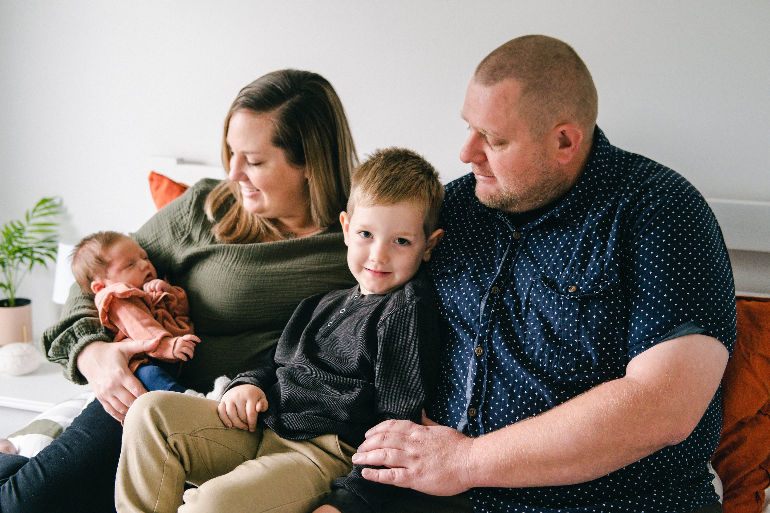 Family, mam dad son and newborn daughter sitting on bed looking at their baby. brother is looking at the camera and smiling. nice natural lit bedroom