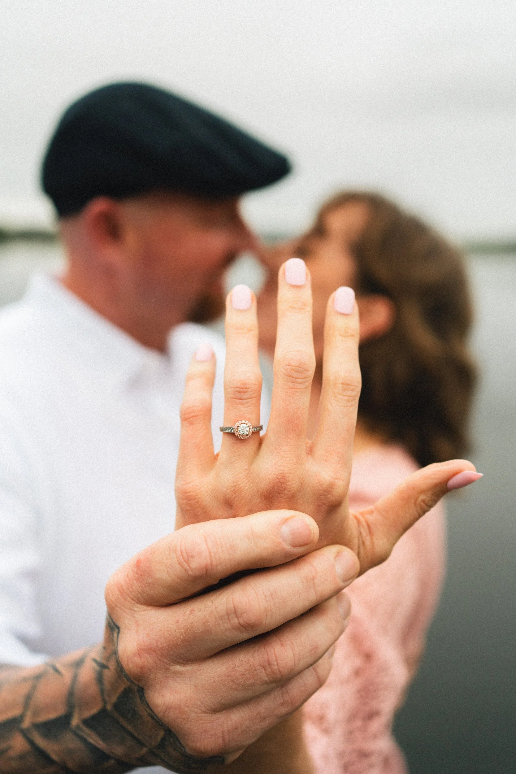 a close up detail image of a stunning engagement ring, one big diamond in the middle, with peach stone around the outside, she is holding out her hand and he is grasping it, they are out of focus in the background touching noses