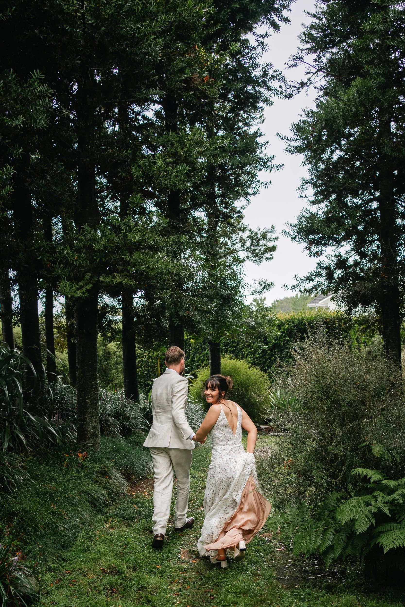 a couple of running away from the camera in a forest of dark green trees, bushes and gress, the bride is wearing a white lace dress with a peach lining,she is looking back over her shoulder smiling, the groom is wearing a cream suit