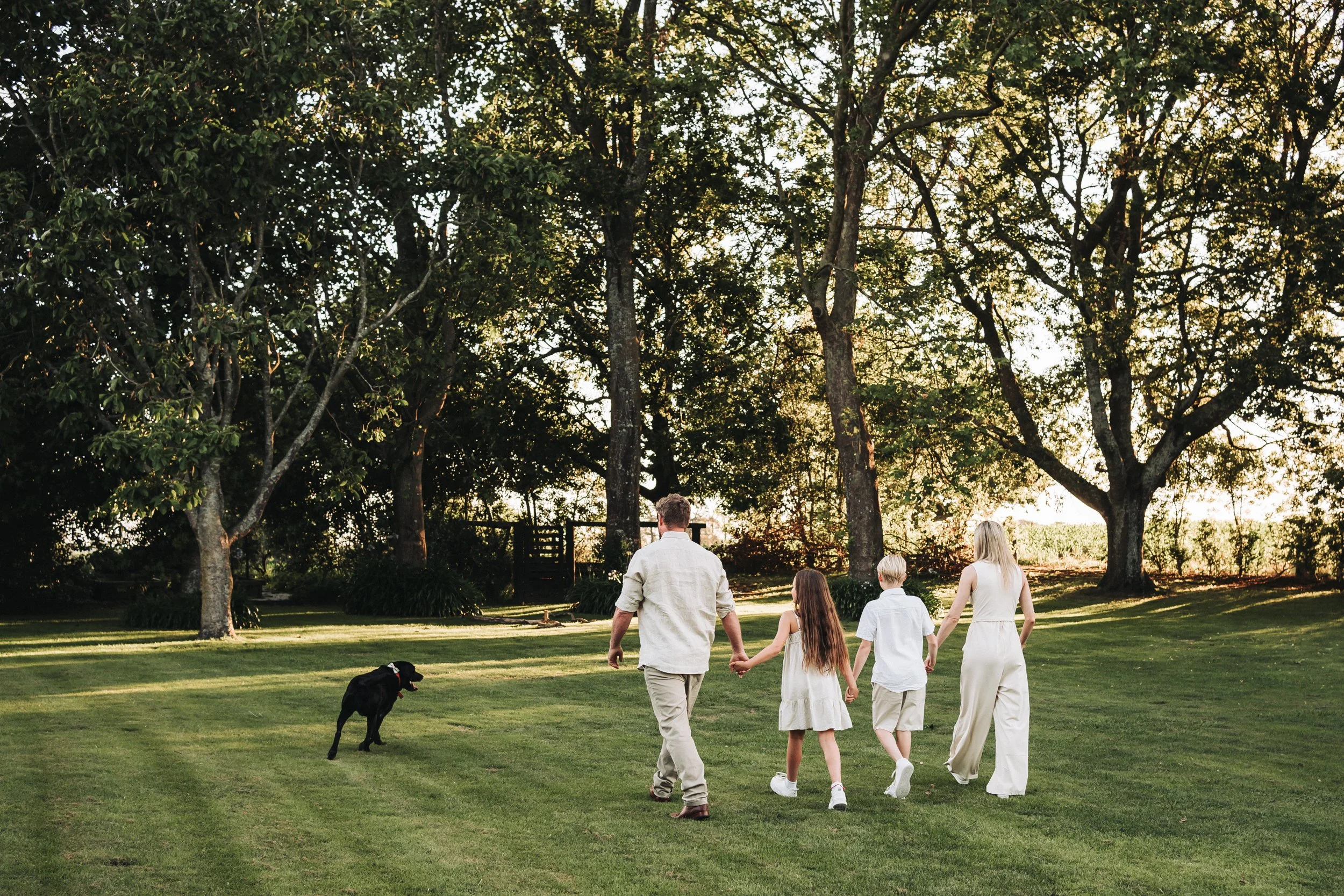 family of 4, all dressed in cream with their black drg, walk hand in hand of into the distance, they are walking on green lawn towards some large trees in the background