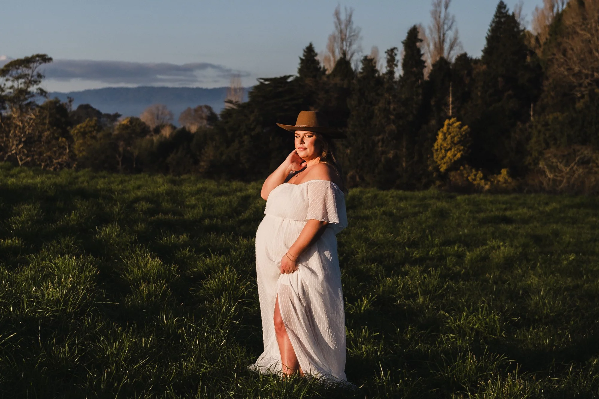 pregnant lady in a floaty white long dress and cowboy hat is standing side on with the setting sun light iluminating her
